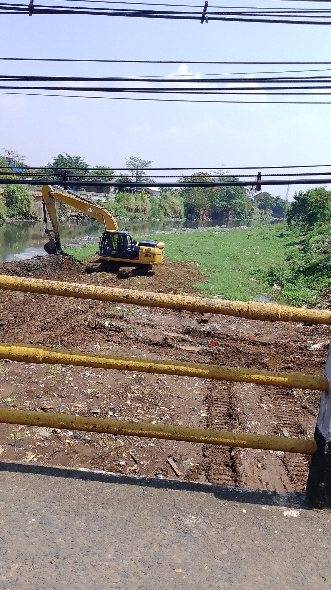 An excavator is parked on a muddy bank next to a river in Baleendah, Indonesia. The river is polluted with visible trash floating on the surface. The bank is covered in mud and debris, with tire tracks from the excavator visible. Beyond the river, there is green vegetation and some buildings. Power lines are strung across the top of the image, obscuring a clear view of the sky. The scene appears to be part of a cleanup or construction project.