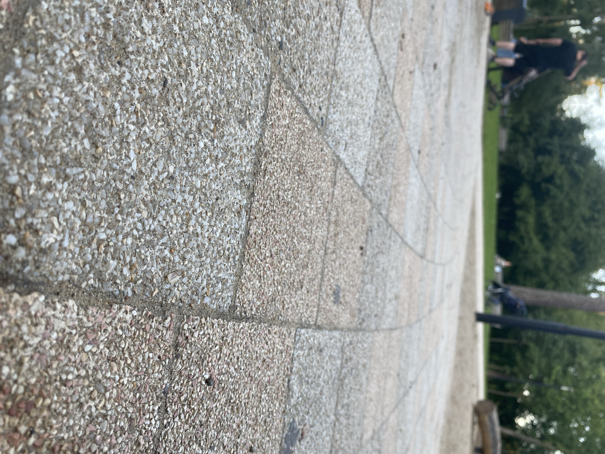 A low-angle shot captures a textured, light-colored paved walkway made of small, interlocked stones, possibly exposed aggregate concrete. The walkway curves gently from the foreground into the background, where a park environment is visible. A person wearing a black t-shirt and shorts stands in the mid-ground, facing away from the camera, near a stroller. In the background, trees with green foliage and a blurred figure can be seen. The scene suggests an outdoor setting, likely a park or public walkway. There is no identifiable information to link this image to Balashikha, Russia.