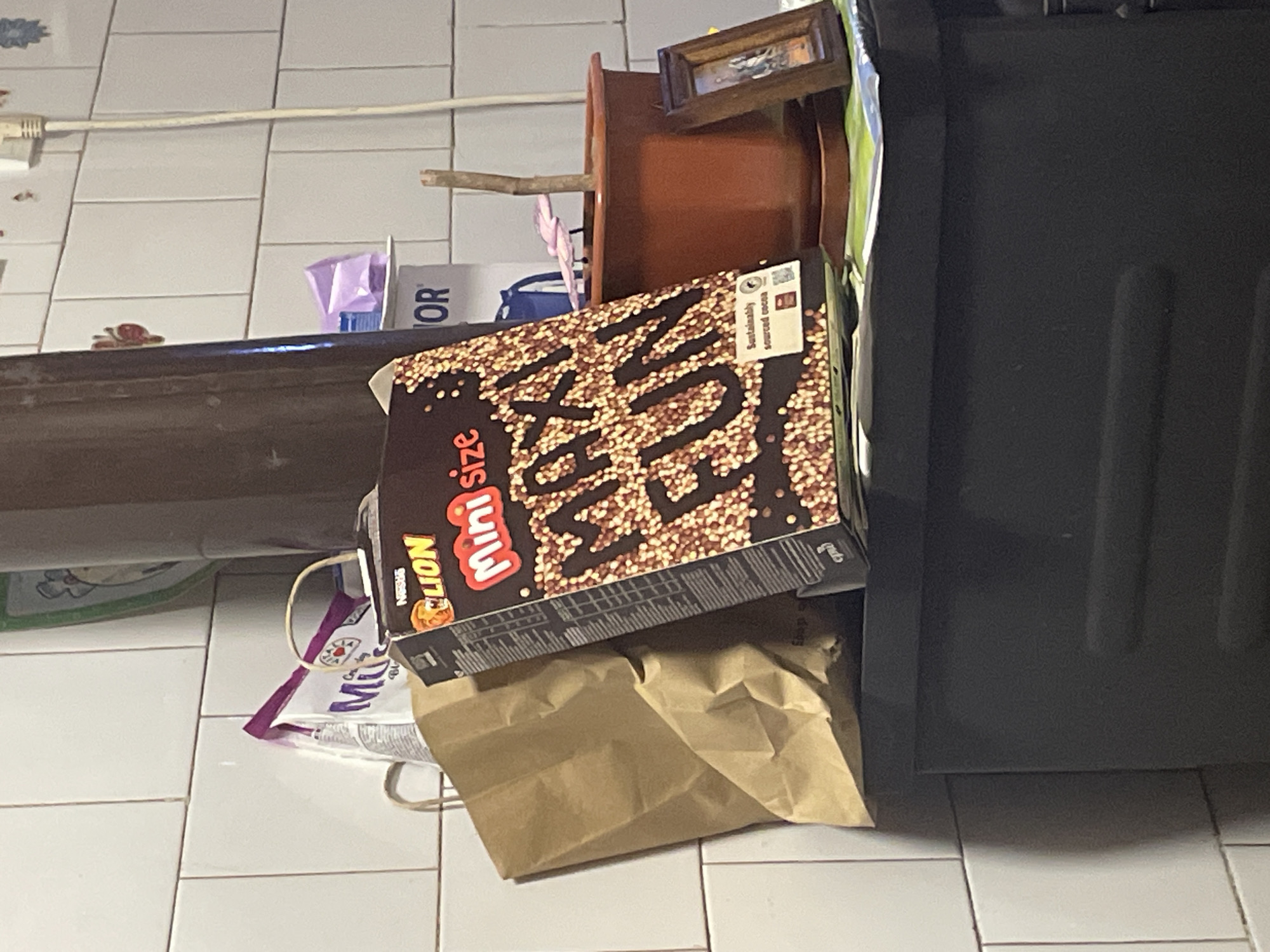 The image displays an indoor scene, likely a kitchen or utility area, in Ihtiman, Bulgaria. The floor is covered with white tiles and light gray grout lines.
In the foreground, a box of cereal is prominently featured. The brand visible on the box is "LION mini size," with the Nestle logo below it. The main product name is stylized as "MINI WEET." The box also carries the text "Sustainably sourced cocoa." Below the cereal box, a crumpled brown paper bag rests on the tiled floor. To the right of the cereal box, a large, dark, rectangular object, possibly a refrigerator or tall cabinet, occupies a significant portion of the right side of the frame.
Behind the cereal box and paper bag, partially obscured, is a white plastic bag with the text "Mil" and a small heart-like logo. Further back, on a surface or shelf, a brown planter pot with a small stick protruding from it is visible. Next to the planter, a small, framed picture or mirror stands upright.
In the background, on the tiled floor, a white electrical cord is plugged into a wall outlet. A small decorative item, possibly a turtle figurine, is also visible on the floor. Other indistinct objects, including a purple item and a box with the letters "OR" on it, are scattered in the background. The lighting suggests an indoor setting, and no specific time of day is discernible. There are no people visible in the image.