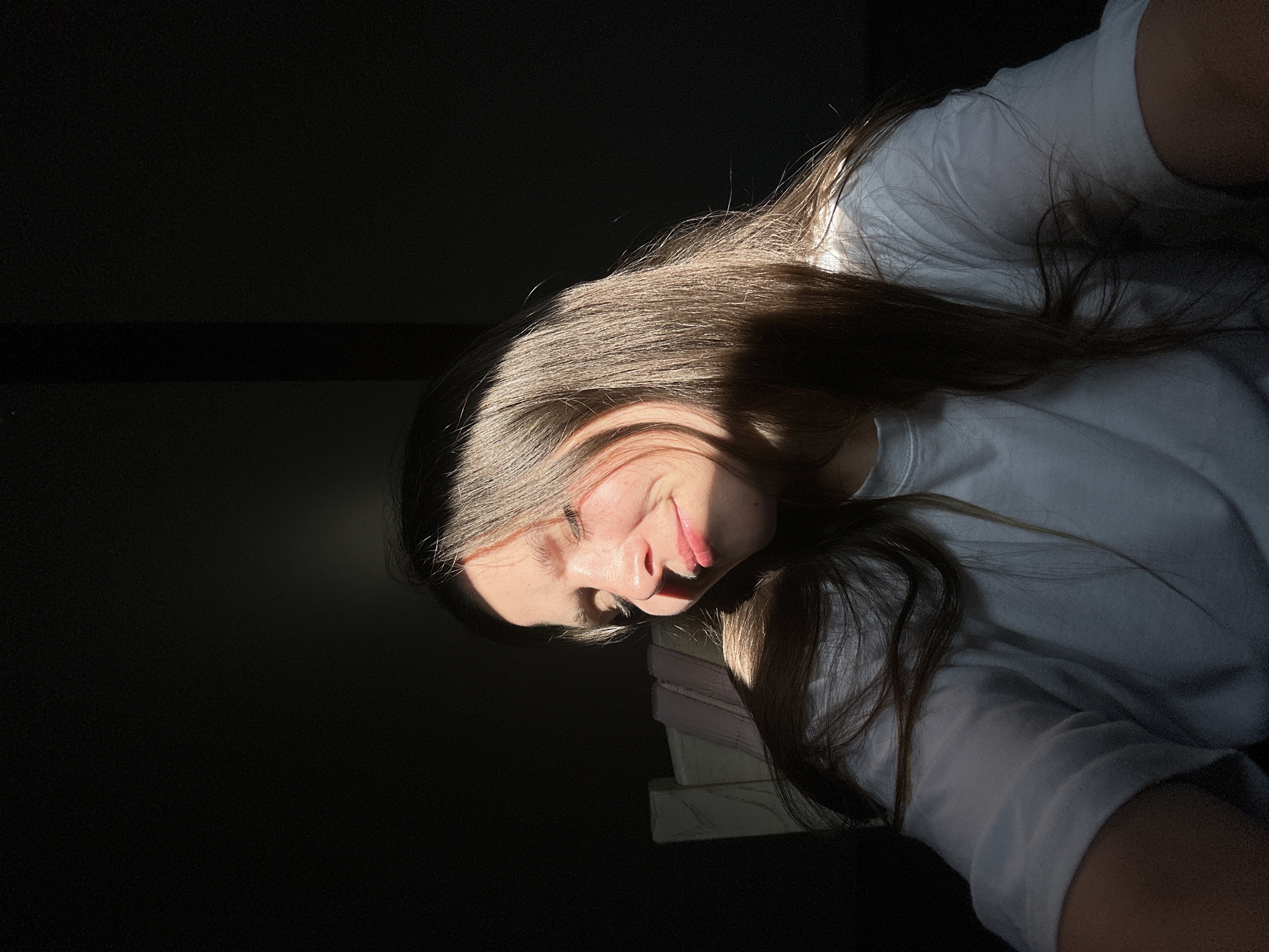 The image shows a young person, likely female, lying down in a low-light indoor setting. The photograph is rotated 90 degrees clockwise, with the person's head positioned on the right side and their body extending towards the left. A strong, directional beam of natural light, presumably sunlight, dramatically illuminates the right side of their face and their long, brown hair, creating stark contrasts with the deep shadows that envelop much of the frame and the left side of their face.
The subject is wearing a white t-shirt. Their expression features a subtle smile or smirk, suggesting contentment or relaxation. Their eyes are open, and their gaze is directed slightly upwards. Their long, flowing brown hair catches the sunlight, highlighting its texture and color. Below their head, a stack of light-colored objects, possibly books or papers, is faintly discernible, serving as a support or part of the surface they are resting on.
The background is predominantly dark, emphasizing the focused illumination on the subject. The quality of light indicates either an early morning or late afternoon setting. There is no visible text present in the image. The overall mood is intimate and tranquil, characterized by the interplay of light and shadow. While the location context is Bartula, Montenegro, no specific geographical features are visible within the image itself.