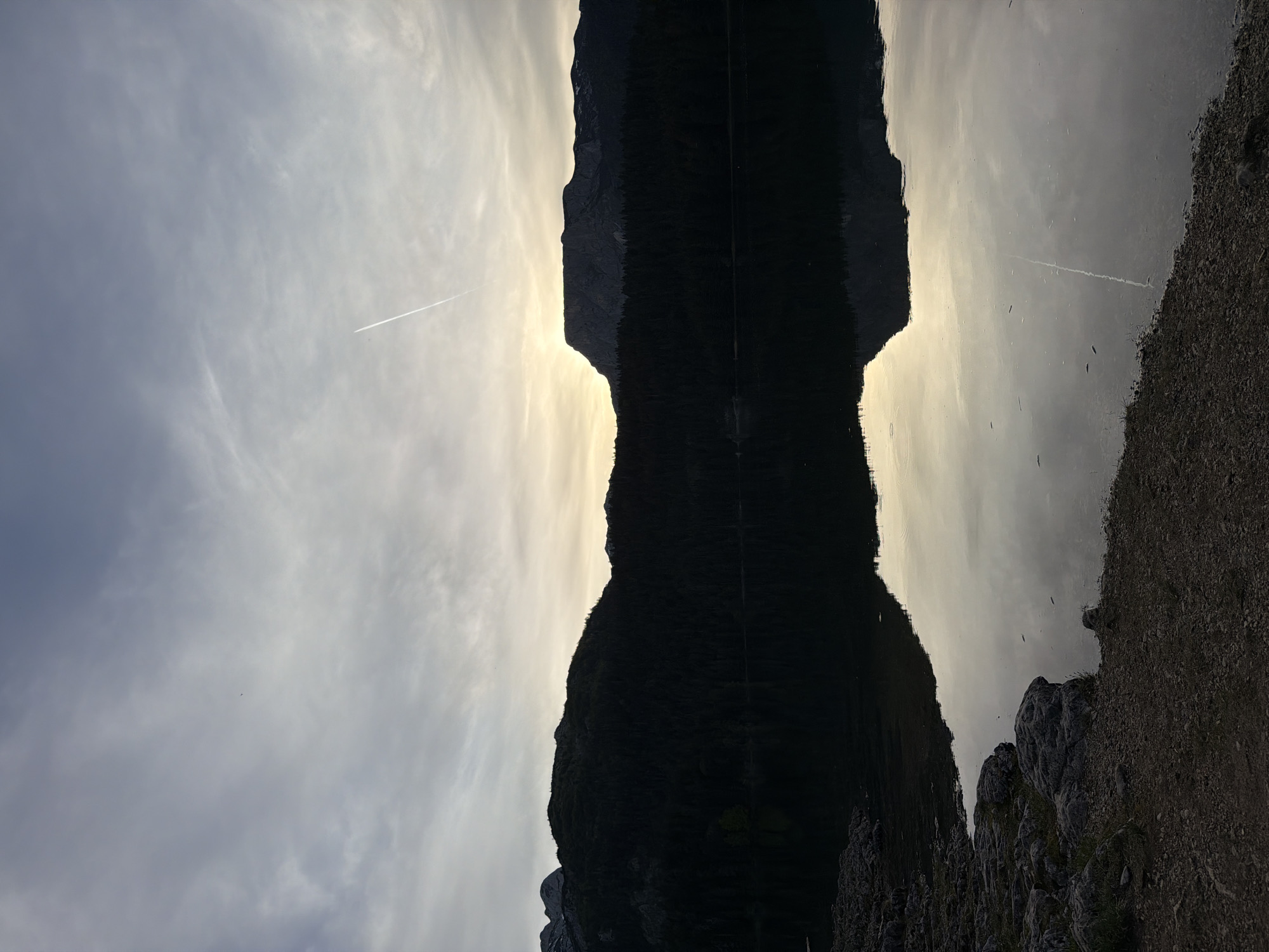The image depicts a mountain reflected in a body of water, likely a lake, in the city of Pitomine, Montenegro. The mountain is mostly in silhouette, with a bright sky behind it, giving the appearance of a dark, inverted shape mirrored in the water. A contrail is visible in the sky, indicating an airplane. The right side of the image shows a rocky shoreline.