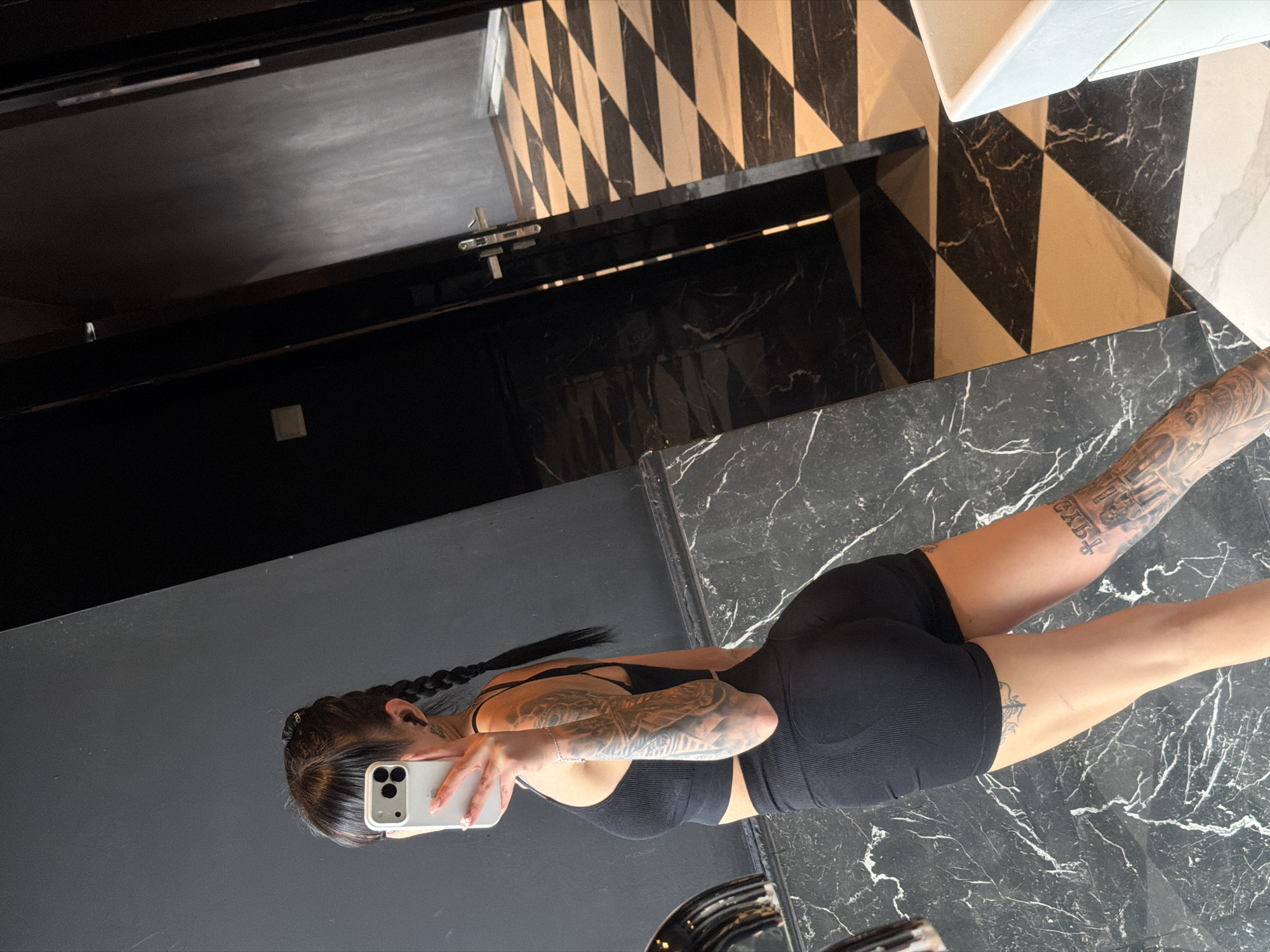 A woman is taking a mirror selfie in a modern bathroom with black and white checkered flooring. She is wearing a black sports bra and black shorts, and has multiple tattoos on her arms and legs. Her hair is styled in two braids. The background shows a dark wall and a door with a silver handle. There is no indication of the location being Plovdiv, Bulgaria.