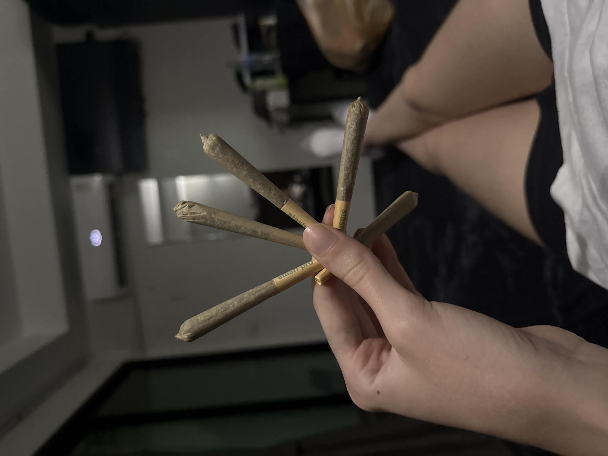 The image shows a person's hand holding five rolled cigarettes, or joints, arranged in a star-like pattern. The person is lying down in what appears to be a bedroom. Legs, clad in dark shorts, are visible in the foreground, suggesting the person is in a relaxed, indoor setting. The background is dimly lit, with a large dark object that could be a television screen mounted on the wall. An air conditioning unit is also visible. The scene suggests a private, domestic environment, possibly within a residential apartment or hotel room. The presence of rolled cigarettes implies recreational use. The overall ambiance is casual and intimate. The location is the city of Bangkok, Thailand.
