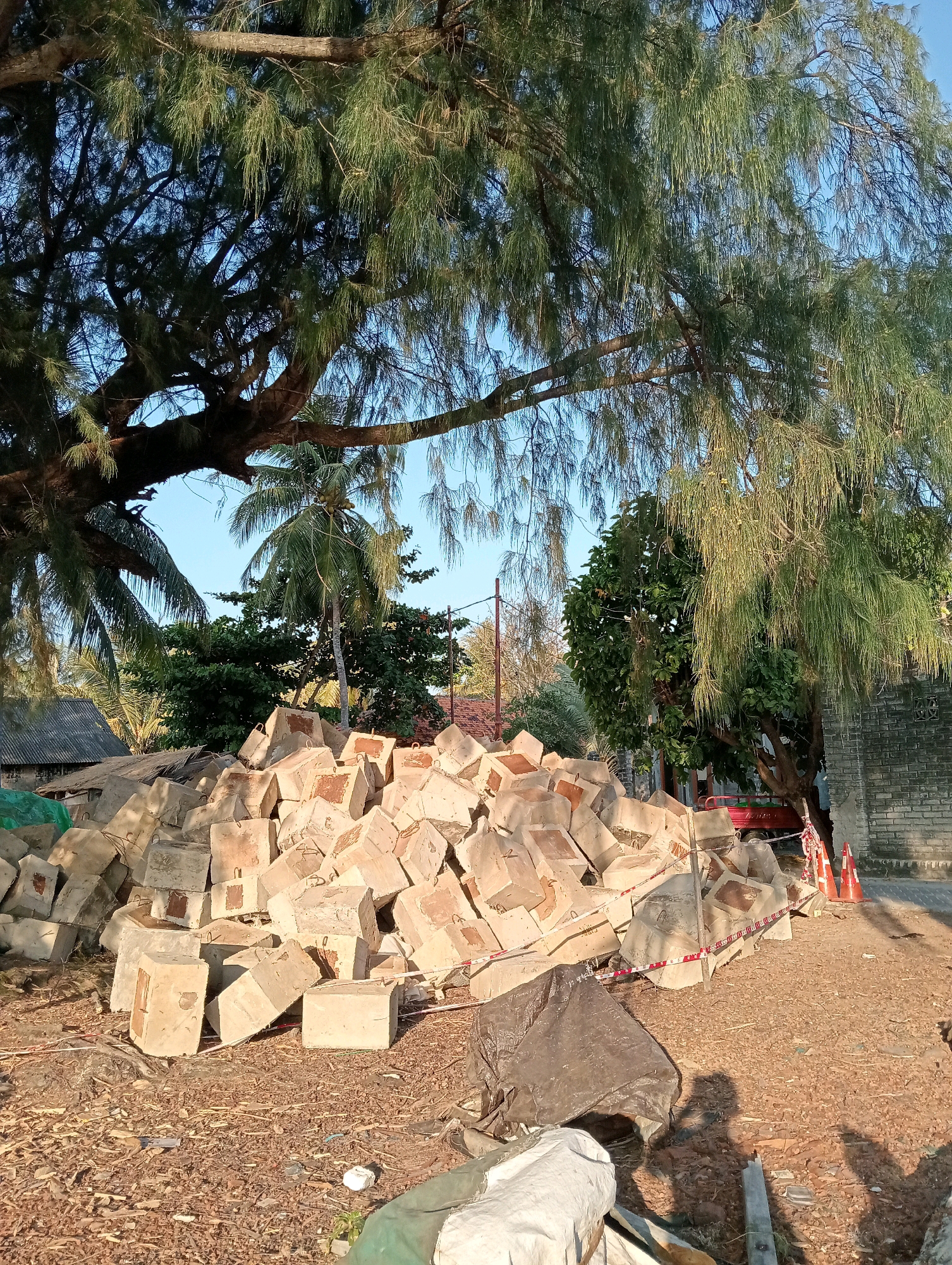 In Pagerungan Kecil, Indonesia, a construction site is depicted with a large pile of concrete blocks, possibly for breakwater construction. The blocks are stacked haphazardly, with some showing metal rings for lifting. A red cart and orange traffic cones are visible in the background, indicating ongoing work or a closed-off area. Surrounding the site are lush green trees, including palm trees, and a clear blue sky. The ground is covered in gravel and fallen leaves. The scene suggests a temporary setup for a construction project in a tropical environment.
