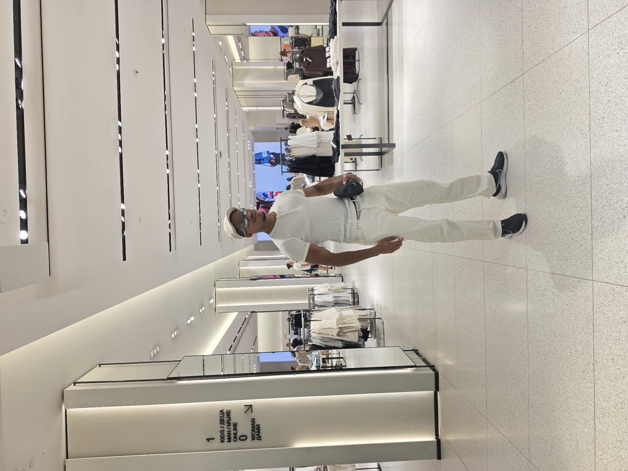 A man, appearing to be in his late 20s or early 30s, stands in a brightly lit, modern retail store. He is wearing a white short-sleeved polo shirt, white pants, a light-colored baseball cap, and sunglasses. He is holding a black pouch in his right hand and is looking slightly off-camera to his right.
The store appears to be a clothing retailer, with racks of clothes displayed on either side of the aisle and tables with folded garments. The flooring is tiled with a speckled white and grey pattern. In the background, large screens display fashion imagery, and other shoppers can be seen browsing.
A prominent sign on a pillar to the left of the man indicates sections for "KIDS / ДЕЦА", "MAN / МЪЖЕ", and "WOMAN / ДАМИ". Based on the visible signage and the modern retail environment, this store is likely located in a commercial district or mall in the city of Varna, Bulgaria.