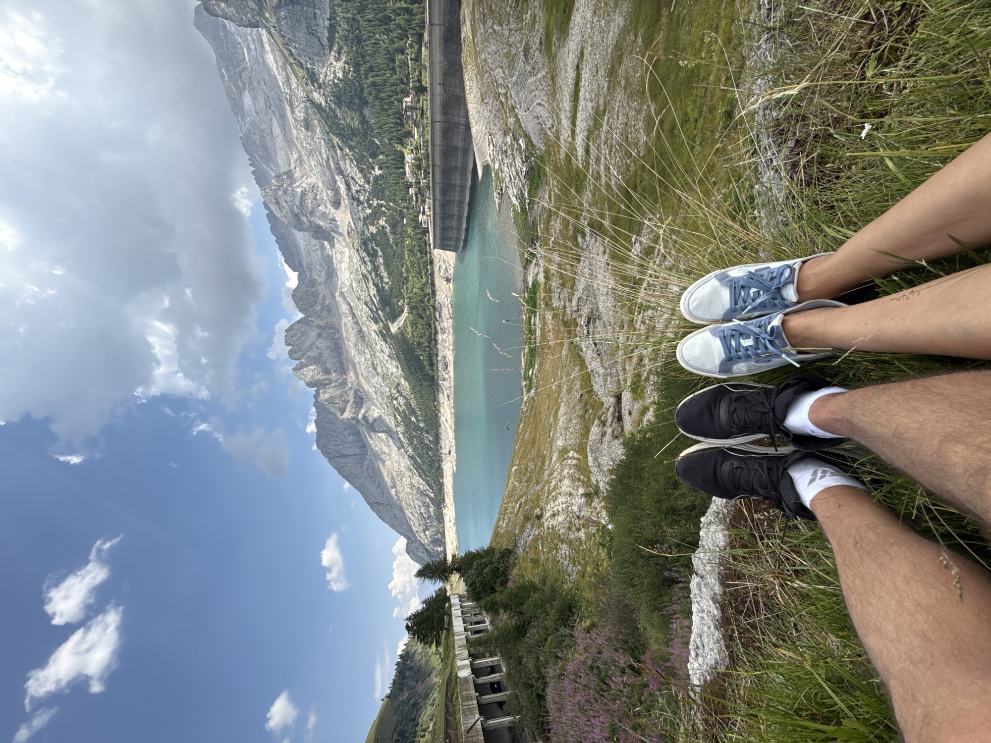 Two individuals are seated in a grassy area, with their legs and feet prominently featured in the foreground. One person wears black athletic shoes with white socks, while the other wears light blue and white sneakers with blue laces. Behind them, a large, turquoise body of water, likely a reservoir, is visible, bordered by a concrete dam. In the background, majestic, rugged mountains rise, their peaks catching the sunlight. The sky is a clear blue, with scattered white clouds. A concrete structure, possibly a bridge or retaining wall, runs along the left side of the frame, lined with trees and vegetation. The overall scene suggests an outdoor recreational setting in a mountainous region. While the image depicts a mountainous landscape, the specific location in the city of Canazei, Italy, cannot be confirmed from the visual information alone.