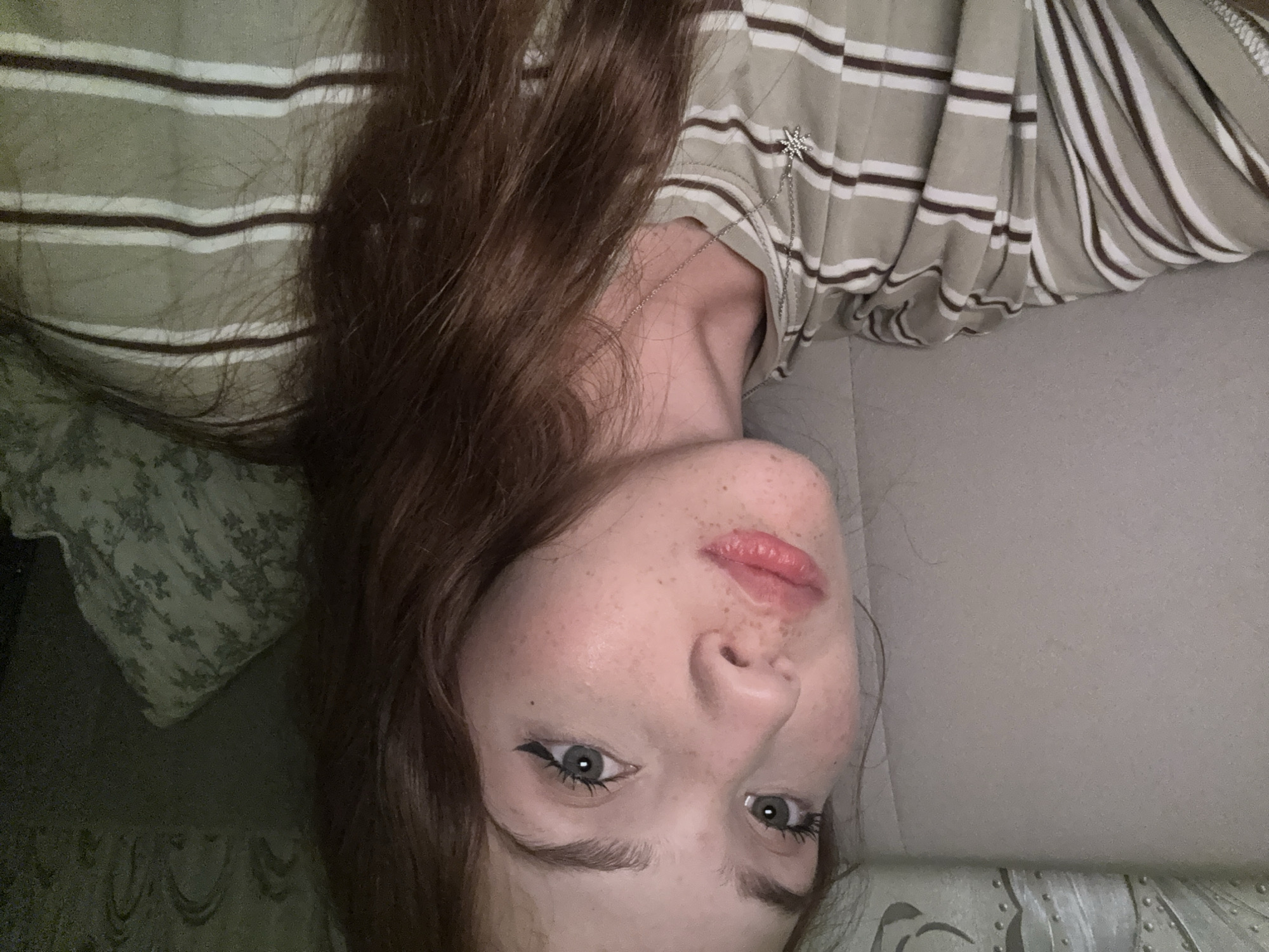A young woman with fair skin, freckles, and auburn hair is captured in a close-up selfie. She is wearing a horizontally striped t-shirt in shades of beige and brown, with a silver pendant necklace. Her eyes are green, and she has on eyeliner. The background appears to be an indoor setting, possibly a bed or sofa, with textured wallpaper or fabric visible. The image is lit with artificial light, creating a warm glow. The location is not specified within the image itself.