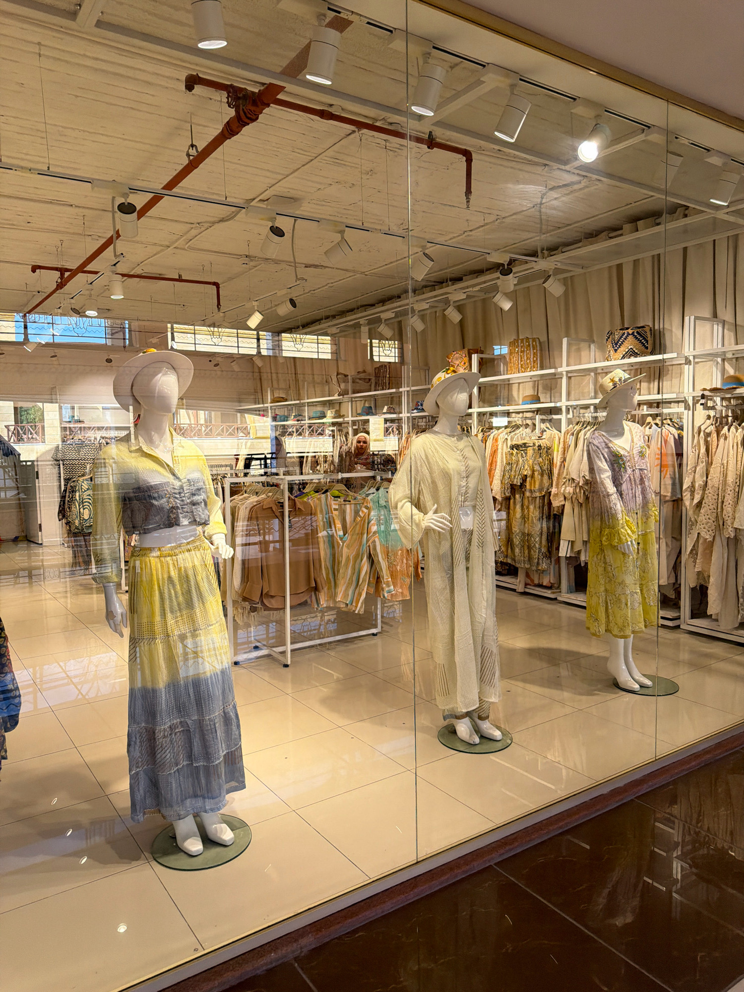 The image shows a retail store window display featuring mannequins dressed in bohemian-style clothing. The store appears to be located in Sharm El Sheikh, Egypt, as indicated by the context. The display showcases three female mannequins, each wearing a long dress or a matching top and skirt ensemble, accessorized with wide-brimmed hats. The clothing is predominantly in light, summery colors like yellow, white, and gray, with some pieces featuring intricate patterns and textured fabrics. Behind the mannequins, racks of similar clothing items are visible, along with shelves displaying handbags and hats. The store's interior is brightly lit with track lighting, and the floor is tiled with a light-colored material. The display is viewed from the exterior through a large glass window, reflecting some of the surrounding environment. A woman in a headscarf can be seen in the background inside the store, browsing the merchandise. The overall scene suggests a boutique selling women's fashion, likely catering to a tourist clientele in a warm climate.