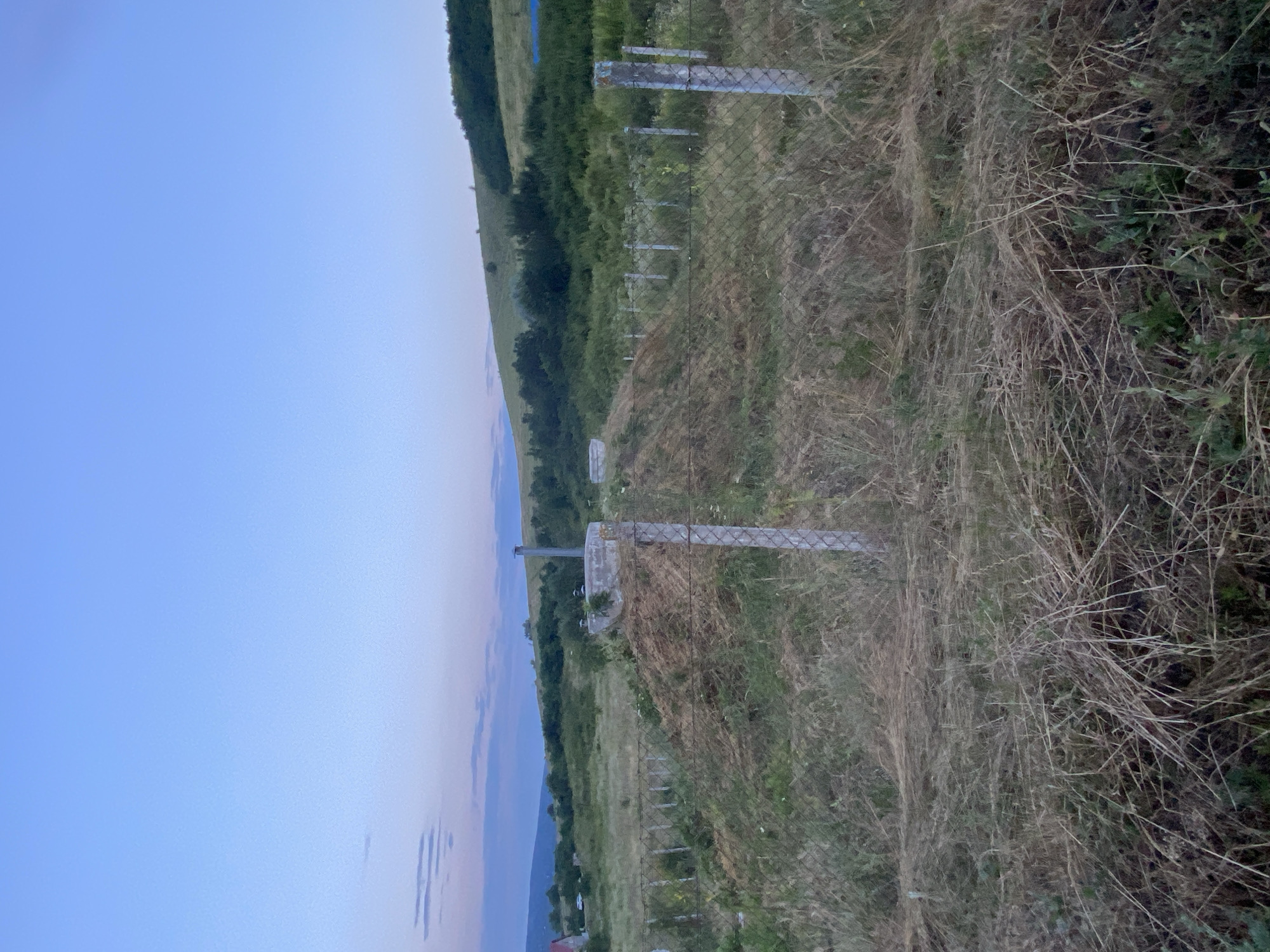 The image depicts an outdoor scene at dusk, characterized by a gradient sky transitioning from light blue to a soft twilight hue with hints of pink and lavender. In the foreground, dry, overgrown grass and hay are visible, interspersed with green vegetation. A chain-link fence with concrete posts runs across the image, indicating a boundary.
Behind the fence, a raised mound of earth, possibly a construction site or a pile of excavated material, is prominent. On top of this mound, there are concrete structures, including a circular one with a metallic pipe extending upwards, possibly a ventilation shaft or chimney. Another smaller rectangular concrete structure is also present.
In the middle ground, the landscape consists of rolling hills covered in dense green vegetation and trees. The terrain slopes upwards towards the right of the frame. A few scattered, thin poles, likely part of a fence or vineyard trellises, are visible in the grassy area.
In the background, a distant hillside with trees borders the horizon. To the far left, a red-roofed building is partially visible, suggesting a rural or semi-rural setting. A white vehicle can be seen in the distance near the building. The overall environment appears to be a developing or undeveloped area, possibly on the outskirts of a town or village, consistent with locations in and around Horodca, Moldova. The lighting suggests the transition from day to night. There are no people or animals clearly discernible in the image.