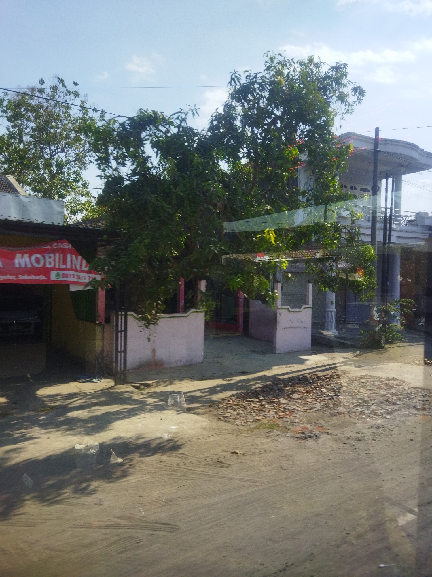 The image depicts a street scene in Nguter, Indonesia, on a bright, clear day. In the foreground, a paved road and dirt area are visible, with shadows cast by trees and structures. Debris, including leaves and small stones, is scattered on the ground. To the left, a red banner with white text reads "MOBILINDO" and displays a phone number, suggesting a business. A car is partially visible inside a covered area. Behind this, a building with a dark roof and light-colored walls stands. Further back, a larger, multi-story building with a white balcony railing and blue accents is prominent, surrounded by lush green trees. The environment appears to be a mix of residential and commercial properties, with a general sense of a suburban or semi-urban setting. There are no people visible in the frame. The overall impression is one of a typical day in a developing town.