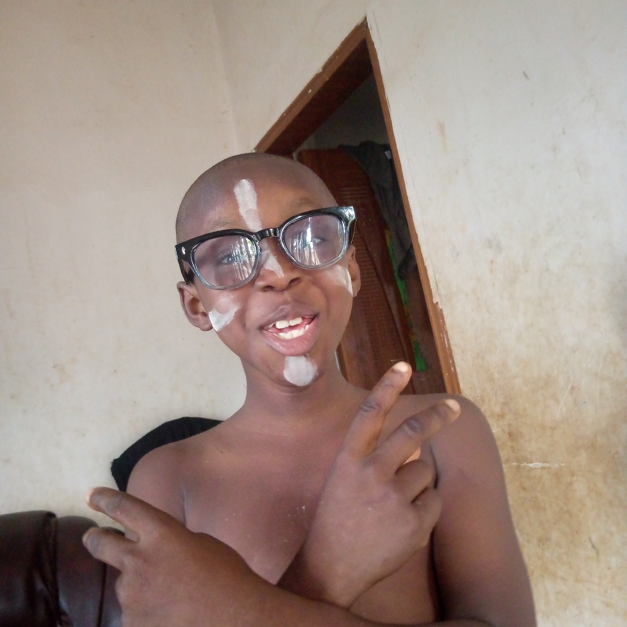 A close-up, chest-up shot captures a young boy with dark skin, a shaved head, and black-rimmed glasses, looking directly at the camera. White marks are visible on his face, including a vertical streak on his forehead, a smear on his right cheek, a patch on his left cheekbone, and a circular mark on his chin. He is smiling widely, revealing his teeth, and appears happy and playful. Both of his hands are raised, making peace signs (V-signs) towards the viewer. He is bare-chested, suggesting an informal setting.
The background consists of light-colored, possibly beige or off-white, walls that show some signs of wear or discoloration. To the boy's right, a wooden doorframe is visible, leading into a darker interior or another room. A sliver of a dark brown or black sofa or cushion is present in the lower left corner of the frame. The lighting is soft and even, suggesting an indoor setting during the daytime. There is no visible text in the image. The overall impression is one of a cheerful and engaging child posing for a photograph.