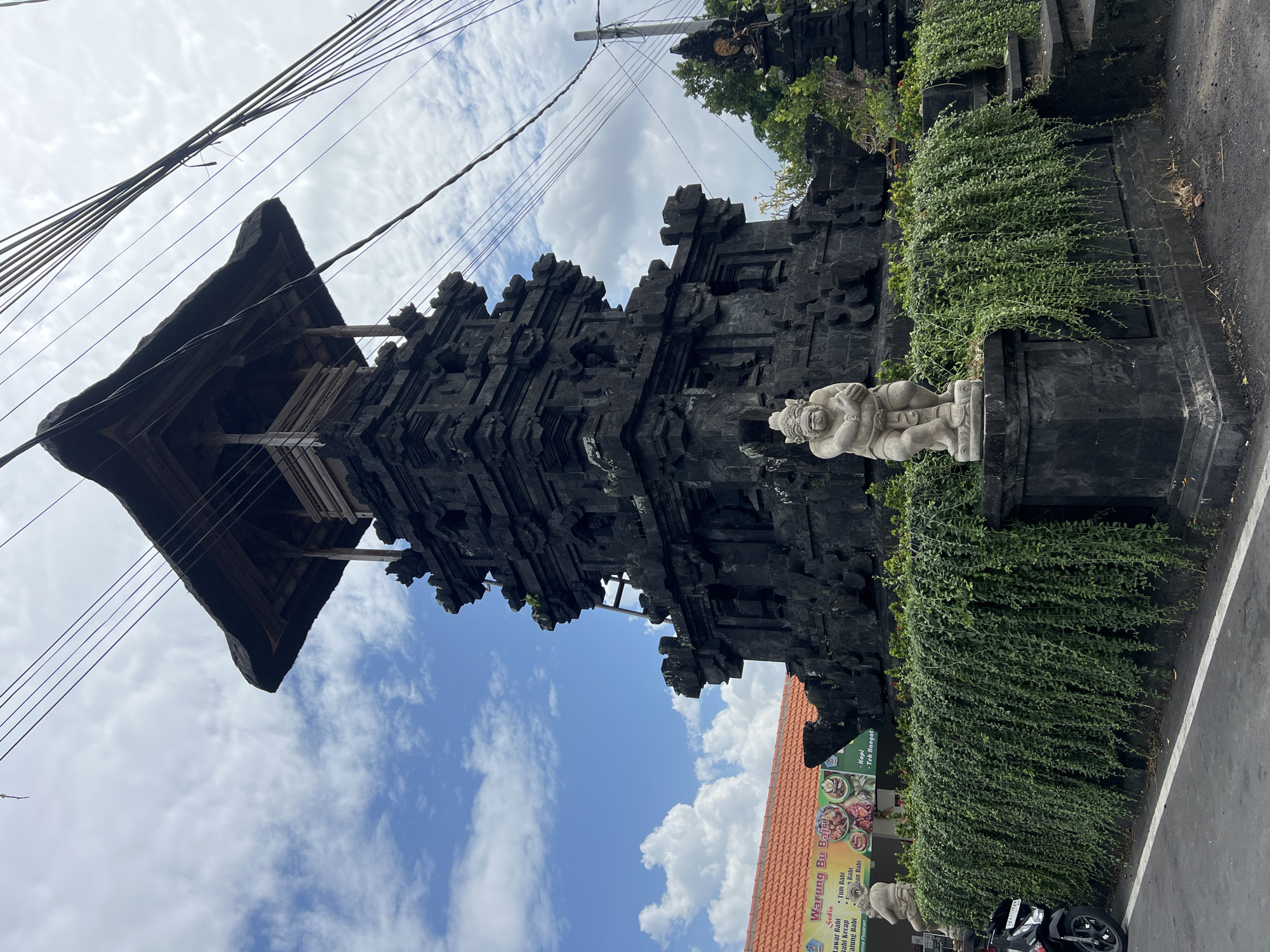 The image shows a stone monument in Sukawati, Indonesia, on a cloudy day. The monument is a multi-tiered structure made of dark stone, with intricate carvings. It is topped with a thatched roof. In front of the monument, there is a statue of a crouching figure made of light-colored stone. The monument is surrounded by lush green vegetation. To the left of the monument, there is a building with an orange tiled roof and a sign that reads "Warung Bu Bajra". A motorcycle is parked to the far left of the image. Overhead, several power lines are visible against the cloudy sky.