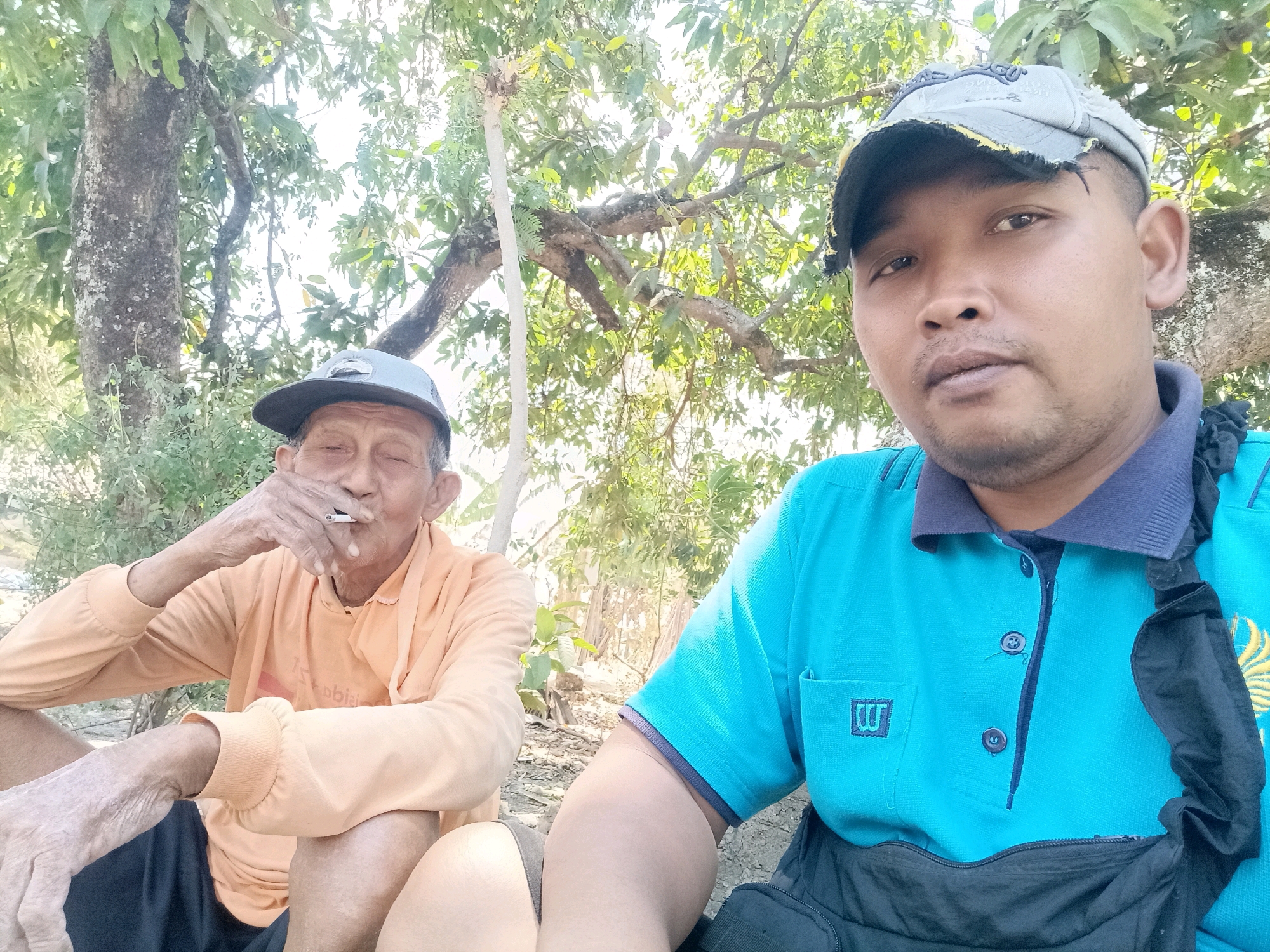 Two men are seated outdoors in what appears to be a rural area, with trees and foliage in the background. The man on the left, wearing an orange long-sleeved shirt and a gray cap, is smoking a cigarette and appears to be older. The man on the right, wearing a blue polo shirt and a gray cap, is closer to the camera and is looking directly at it. The environment suggests a warm, possibly sunny day. The presence of trees and the ground texture indicates a natural setting, potentially outside a village or on the outskirts of a town. The image captures a candid moment between the two individuals. There is no visual information to specifically place the scene in Sumber Bendo, Indonesia.