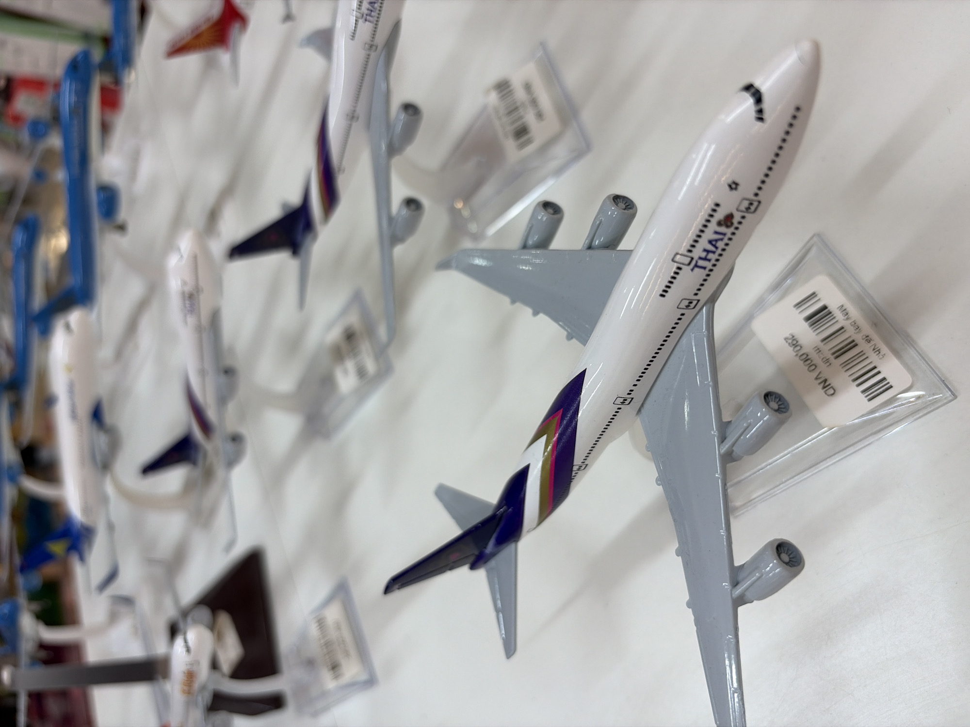 The image displays a collection of model airplanes, primarily from the airline Thai Airways, arranged on a white surface. The foreground features a detailed model of a Thai Airways jet with its engines, wings, and tailfin clearly visible. The tailfin is painted with purple, white, and gold stripes. Attached to the model is a price tag or information card with Vietnamese text and a barcode. In the background, several other model airplanes are visible, some with blue livery and others with red. The lighting suggests an indoor setting, possibly a retail store or a display area. Given the presence of Vietnamese text on the price tag, it is plausible that this scene is located in a city in Vietnam, such as Đà Nẵng, where such merchandise might be sold. No people are visible in the image.