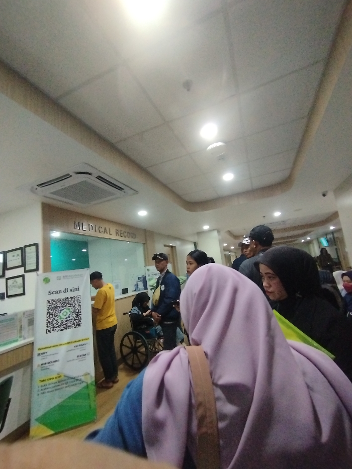The scene takes place inside the "Medical Record" section of a hospital in Soreang, Indonesia. The location is identified by the coordinates -7.032 latitude and 107.527 longitude. The scene is a crowded waiting area with a group of people waiting for their turn. There are four people standing, including a man in a yellow shirt, a man in a blue shirt, and a woman in a pink headscarf and a woman in a black headscarf. The man in the yellow shirt is standing near a large sign with a QR code on it, which reads "Scan di sini."  A person sits in a wheelchair facing the wall. The ceiling has several recessed light fixtures and a white tiled ceiling.