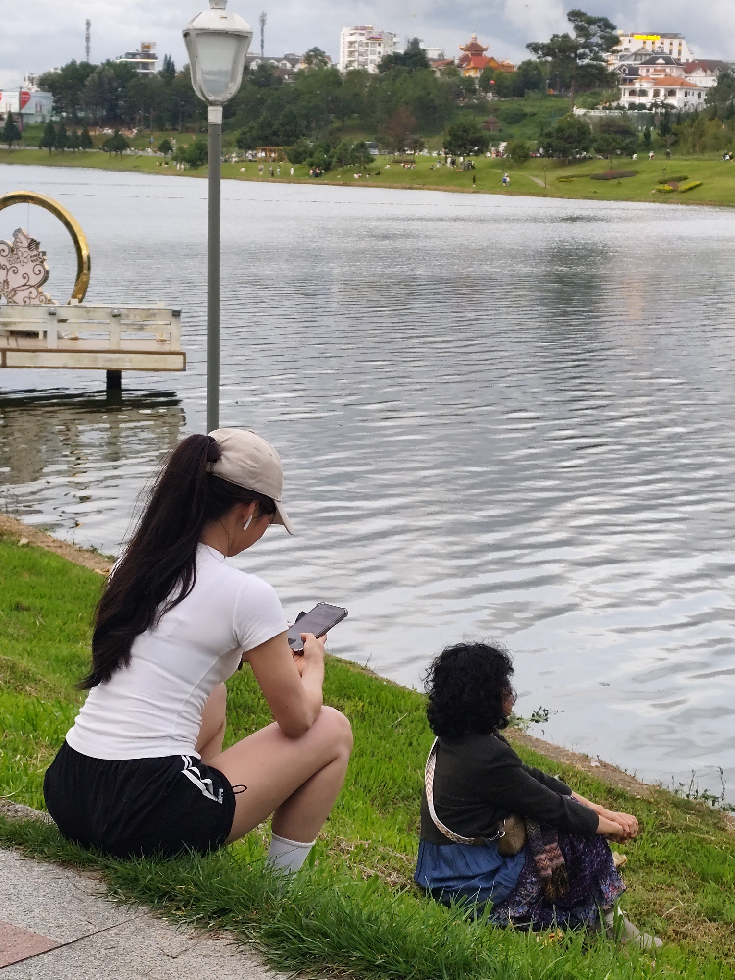 In the city of Xuân Hương - Đà Lạt, Vietnam, two individuals are observed near the lake. In the foreground, a woman wearing a white t-shirt, black shorts with white stripes, white socks, and a beige baseball cap is squatting on the grass, holding a smartphone and looking down at it. She has her hair pulled back in a ponytail and appears to be wearing earbuds. To her right, another individual, with curly dark hair, is seated on the grass facing the lake. They are wearing a dark jacket over a blue skirt and a patterned scarf draped over their shoulder with a small bag. The background features the rippling surface of Xuân Hương Lake, with a lamppost and decorative elements to the left. Further in the distance, across the lake, a tree-lined hillside is visible with buildings and structures dotting the landscape under an overcast sky.