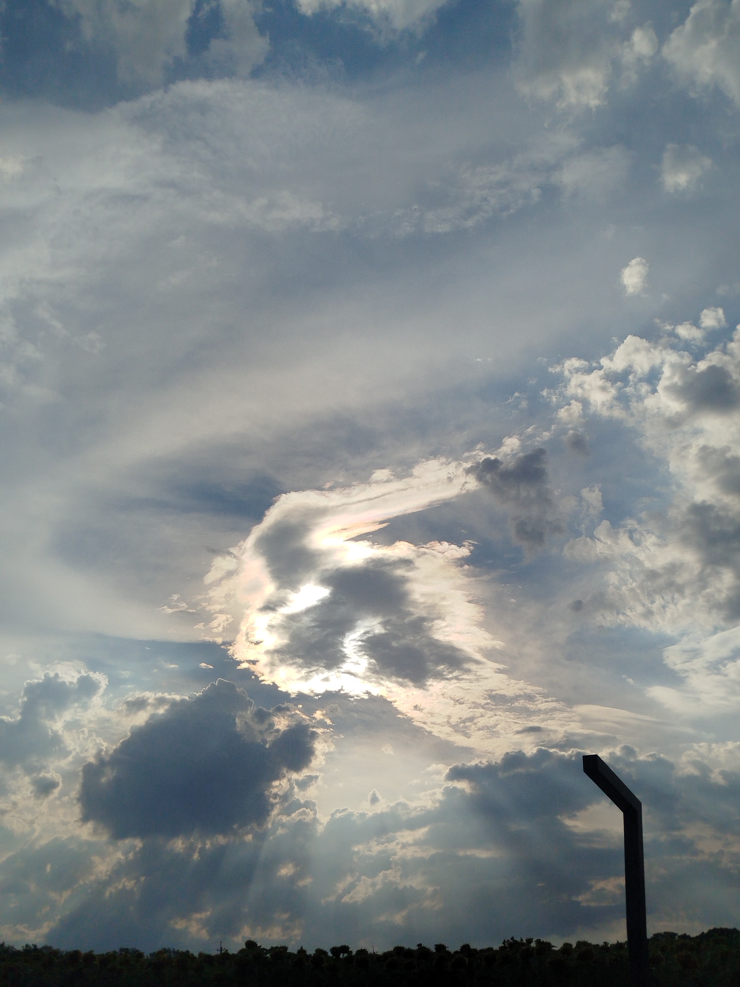 This is a photograph of a cloudy sky with some sunlight breaking through. The sun is partially obscured by clouds, creating a dramatic lighting effect with rays of light beaming down. There is a dark, angular object in the foreground, silhouetted against the sky. Below the sky is a dark band of trees. The image does not contain any people or specific urban landmarks that would allow for identification of a location within Verkhn'odniprovs'k, Ukraine.