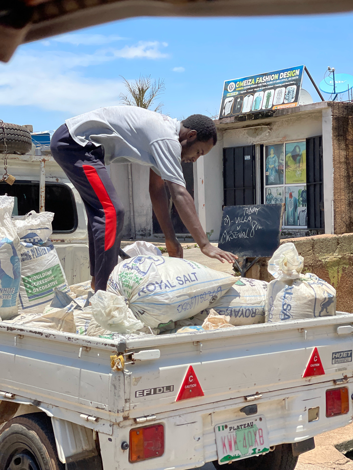 A young Black man, wearing a grey t-shirt and dark sweatpants with a red stripe down the side, bends over a white flatbed truck filled with large bags of what appears to be salt and plaster. He is reaching towards a blackboard propped up amongst the bags. The blackboard has writing on it in white chalk, indicating "TODAY 20-VILLA CRONAV 8PM". Behind the truck is a building with a sign reading "OMEIZA FASHION DESIGN" and several framed fashion advertisements displayed in a window. The scene is outdoors under a bright, partly cloudy sky, suggesting a warm day. The truck has a license plate that reads "PLATEAU KWK 40XB". The overall setting appears to be a commercial or residential area in the city of Jos, Nigeria, given the visible signage and license plate.