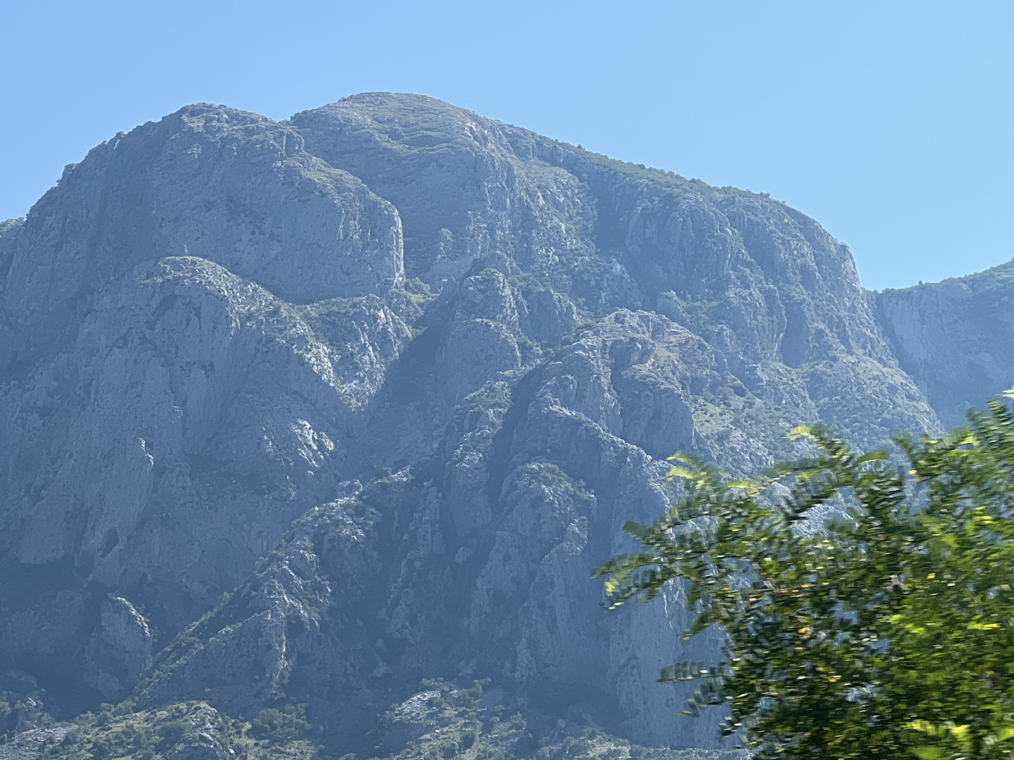 The image depicts a vast, imposing mountain range under a clear, bright blue sky, suggesting a sunny day. The mountains are rugged and rocky, with steep inclines and deep crevices. Patches of green vegetation cling to the slopes in places, hinting at a somewhat arid climate. In the foreground, a tree with vibrant green leaves is blurred due to motion, indicating the photograph was likely taken from a moving vehicle. The sunlight casts shadows on the mountains, highlighting their dramatic contours and textures. The overall scene evokes a sense of awe and the grandeur of nature. There are no visible people, objects, or text in the image. The setting is mountainous, and given the context of Škaljari, Montenegro, it is likely a view of the Lovćen mountain massif or a similar prominent peak in the region. The clear sky and bright sunlight suggest it is daytime, likely late morning or early afternoon.