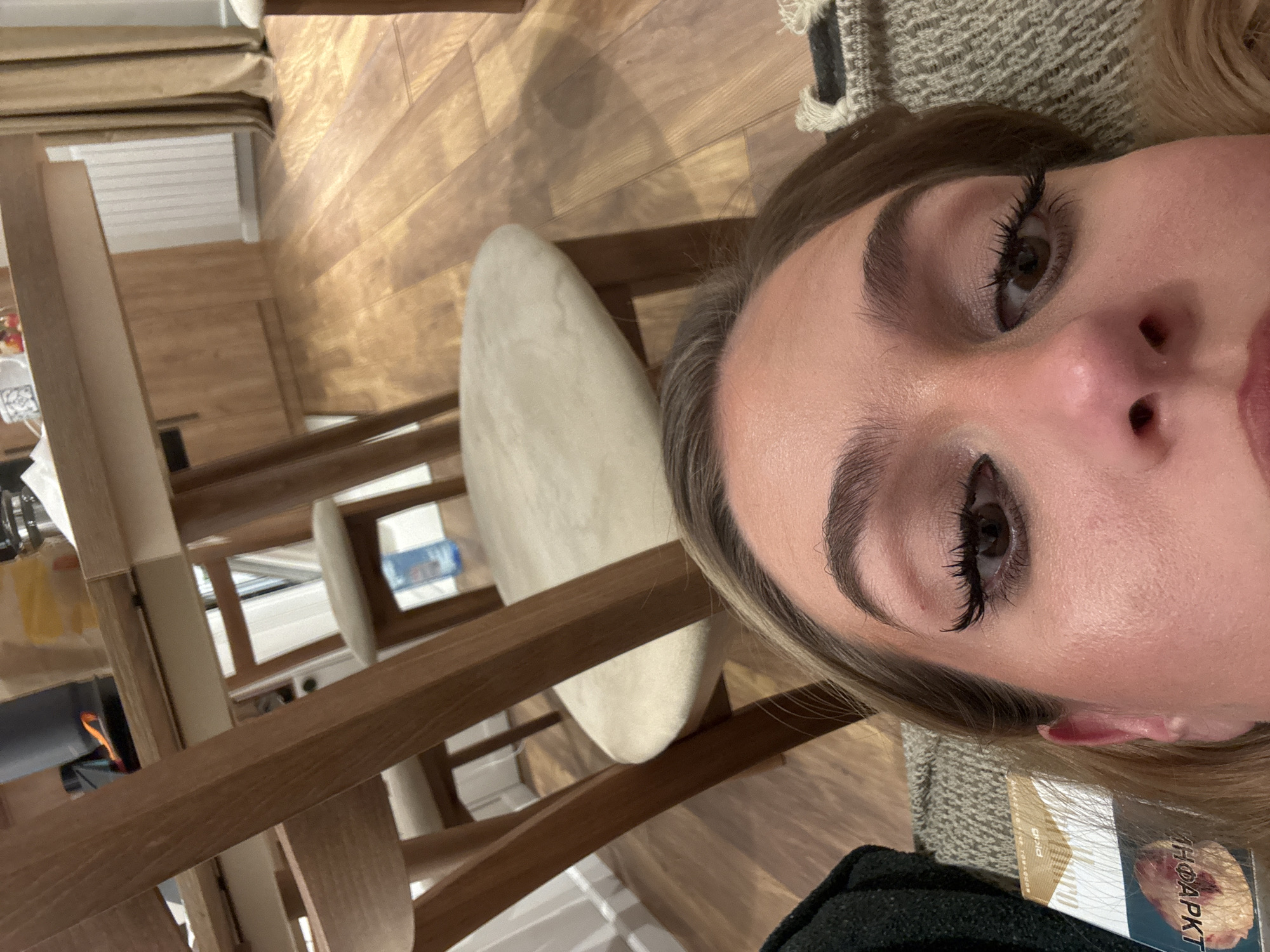 A close-up, rotated image shows a person's face, primarily the right side, from a high-angle perspective. The person, appearing to be female, has well-defined eyebrows and long, dark eyelashes, suggesting the use of mascara or extensions. Their skin appears smooth, with subtle hints of makeup like blush. Light brown or blonde hair frames the face, and a dark-colored garment, possibly a sweater or jacket, is visible on their shoulder, along with a textured, light-colored blanket or throw.
The background reveals an indoor setting with light-toned wooden flooring that has a distinct grain. To the left, a wooden dining table and chairs with light-colored, possibly beige or cream, upholstered seats are visible. Through the chair legs, a glimpse of a light-colored wall or baseboard and what seems to be a window or light source can be seen. On the table, there are indistinct items that appear to be a white napkin and some colorful objects.
The overall lighting suggests an indoor environment. On a rectangular object partially obscured near the person's right shoulder, the words "Gold" and "THOAPKT" (or a similar arrangement of letters) are visible, along with fragmented text that might include "Bia" or "Bid". The image is factually a close-up, likely a selfie, with the person looking towards the camera.
