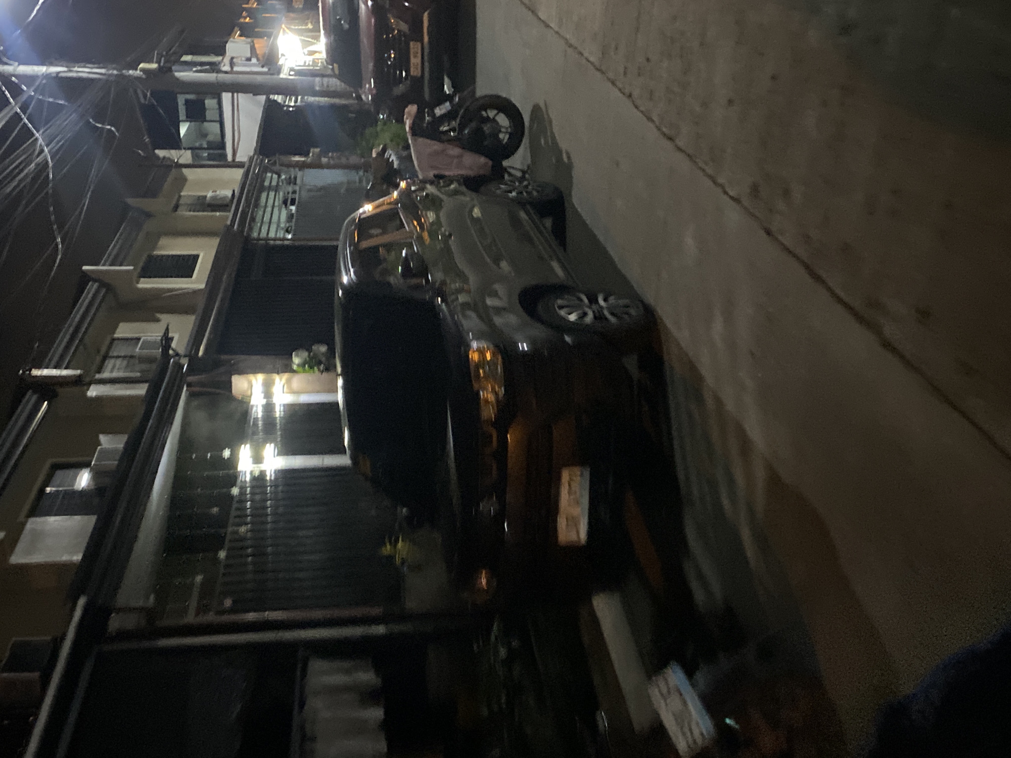 The image depicts a nighttime street scene in a residential or mixed-use area of Davao City, Philippines.
In the foreground, a dark-colored Suzuki S-Presso compact SUV is parked on the right side of the asphalt road, facing away from the viewer. Its headlights and amber signal lights are visible. A black motorcycle, appearing to be a scooter, is parked upright next to the S-Presso, leaning slightly towards its right side, with a light-colored (possibly pink or beige) cloth draped over its seat. The motorcycle's rear wheel is visible on the sidewalk.
Further down the street on the right, a red pickup truck, possibly a Nissan Navara or similar model, is parked. Its front license plate, partially visible, reads "XJG 903".
The street is lined with multiple two-story concrete buildings, characteristic of residential structures in the Philippines. These buildings feature windows, some with security bars, and multiple air conditioning units. The building closest on the left has a dark-colored roll-down gate or solid wall at street level. Overhead, a dense network of utility wires runs across the street, supported by a prominent utility pole on the right side of the frame. A concrete sidewalk parallels the road.
Artificial light sources, likely streetlights, illuminate the scene, casting prominent shadows from the vehicles and utility pole. On the far left foreground, some discarded items or refuse are visible on the ground. No individuals are present in the image. The scene is static, showing parked vehicles and residential buildings under artificial illumination.