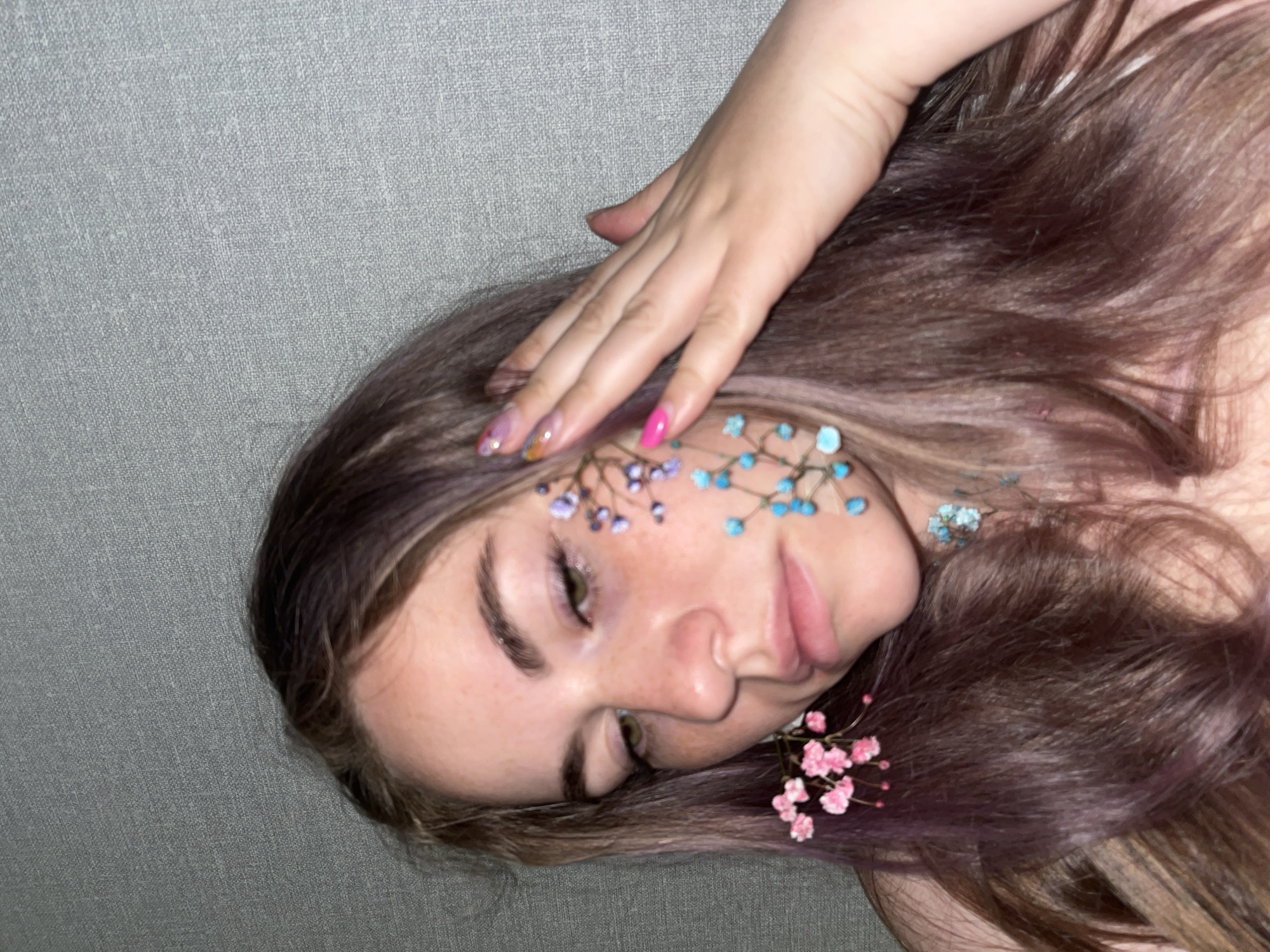 A woman with long brown hair lies on a gray surface, possibly fabric, with her eyes closed. Small blue and purple flowers are placed on her face near her forehead and cheek, and pink flowers are near her neck. Her right hand rests on her hair. She has painted fingernails, with one nail a bright pink color and others with a multicolor design. The background appears to be a simple, textured gray surface. There is no information available in the image to determine its location in the city of Chaikovsky, Russia.
