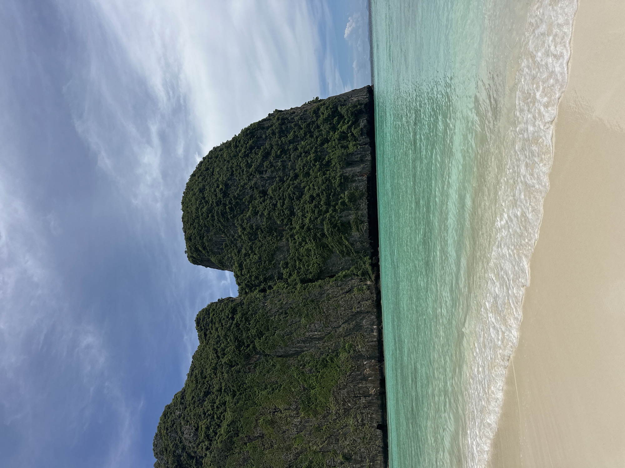 This media file captures a serene tropical beach scene in Ao Nang, Thailand. The image is oriented vertically, showcasing a dramatic limestone karst cliff covered in lush green vegetation. The cliff face dominates the left side of the frame, its rugged texture contrasted with the vibrant foliage. To the right, the clear, turquoise water of the Andaman Sea gently laps against a pristine, sandy shore. Wispy white clouds drift across a bright blue sky, suggesting a pleasant, sunny day, likely mid-morning or early afternoon given the lighting. The overall mood is tranquil and idyllic, conveying a sense of natural beauty and peacefulness. There are no people or objects visible in the image, focusing solely on the natural landscape. No text is present.