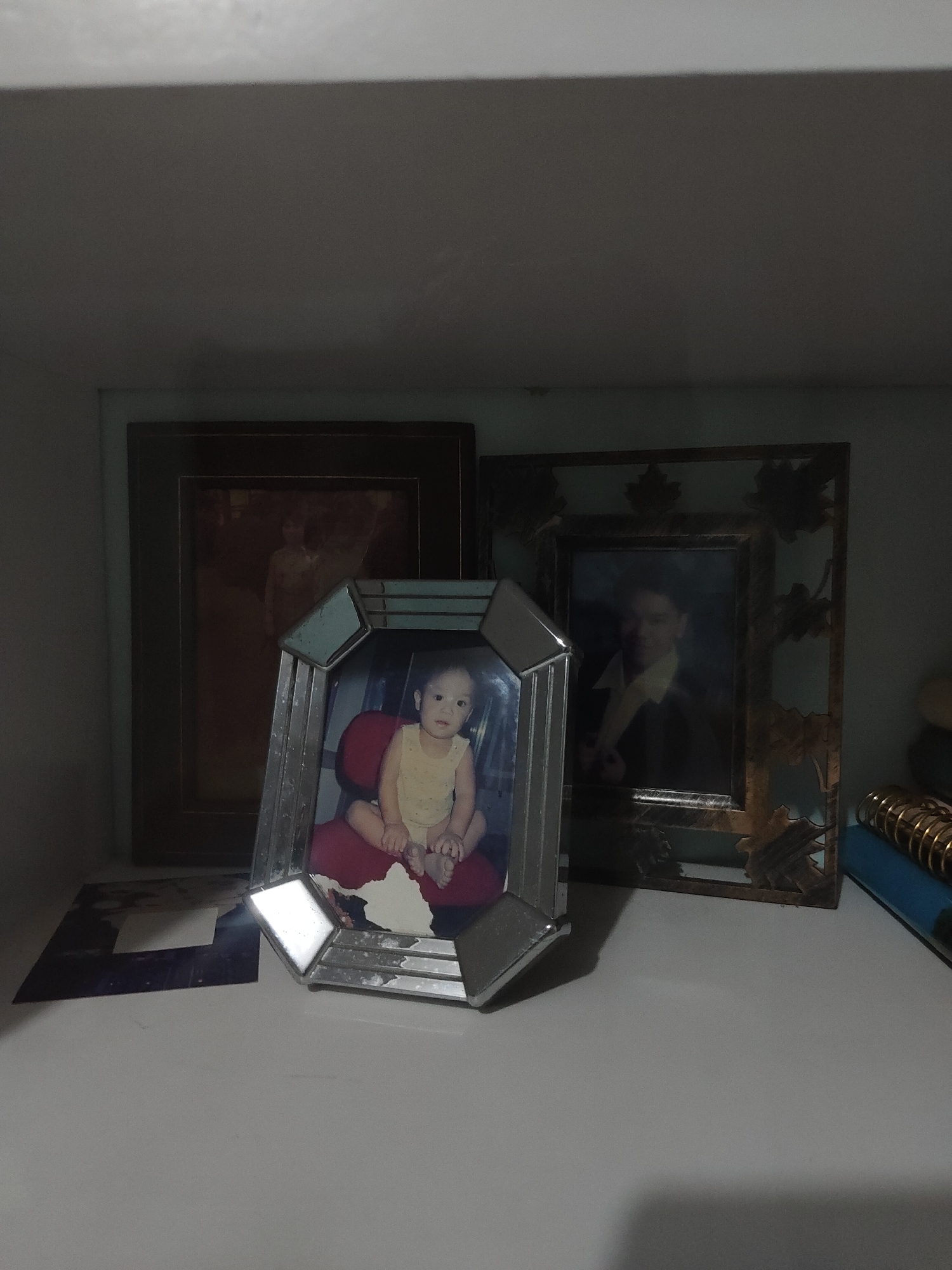 A photograph captures a collection of three framed images displayed on a light-colored shelf or surface within an interior setting in the city of Unknown, Unknown. The scene is illuminated by ambient light, casting soft shadows across the objects.
On the left, a vertically oriented dark brown wooden picture frame contains a faded, sepia-toned photograph of a person, likely an adult female, with indistinct features. Centrally positioned and slightly forward, an octagonal silver-toned picture frame with faceted edges shows signs of wear and tarnishing. This frame holds a color photograph of a light-skinned infant or toddler with dark hair, wearing a light yellow top, seated on a red cushion and looking directly at the viewer. To the right, a rectangular picture frame with an ornate, possibly metallic, leaf-patterned border in a bronze or gold hue surrounds a picture with a dark blue or green inner border. Inside is a color photograph of a young man, appearing to be in his late teens or early twenties, with dark hair, dressed in a dark jacket over a prominent white or cream-colored collared shirt, also looking towards the viewer.
Beneath the central silver frame, a dark, flat object, possibly an unframed photograph or paper, lies on the surface, partially obscured by a smaller, white rectangular object. On the far right, a segment of a spiral-bound notebook or planner with gold rings is visible, resting atop a blue book or binder. The background wall behind the frames appears to be a muted light green or blue, while the upper portion of the image displays a plain light grey surface.