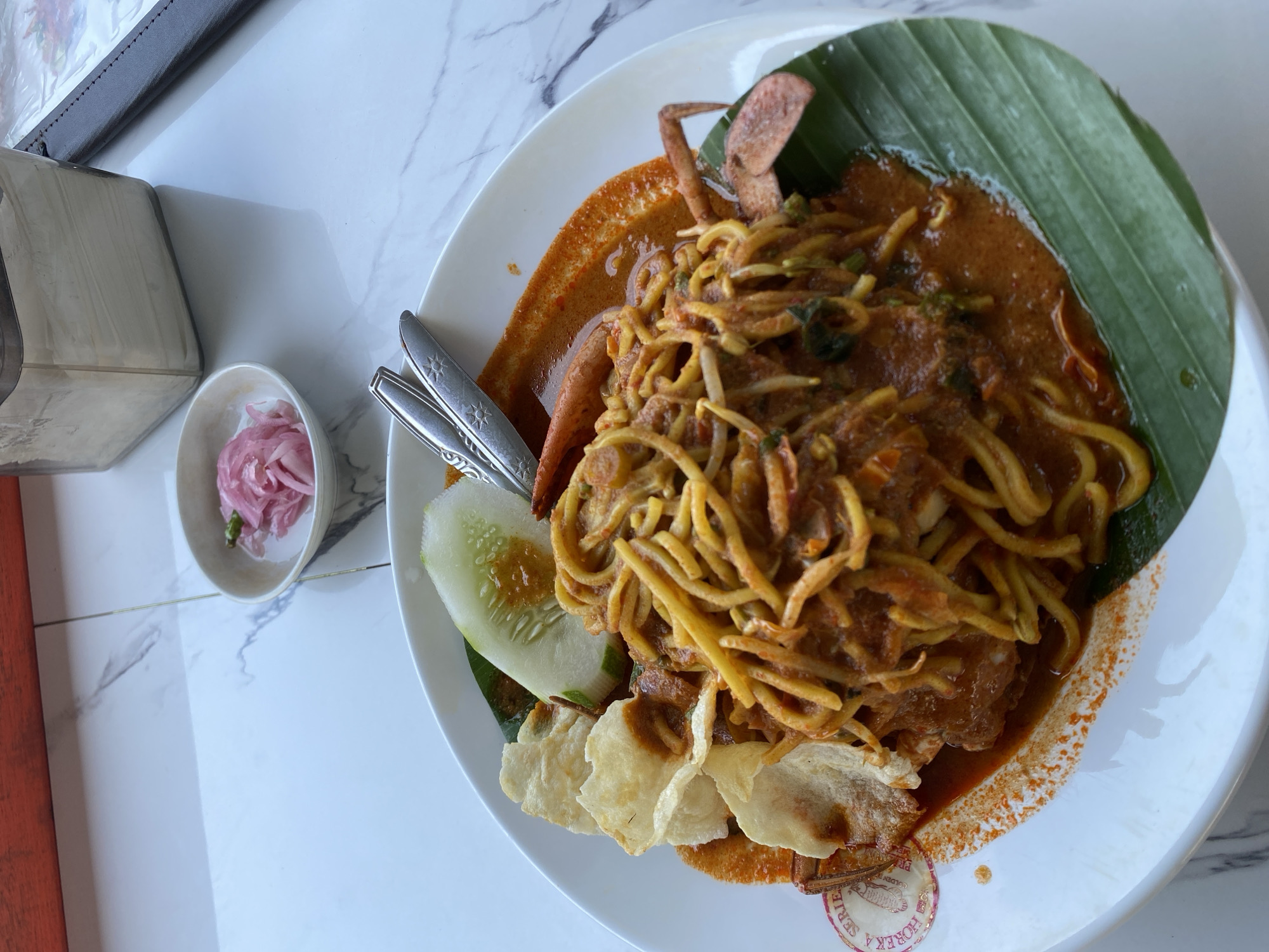 A close-up, eye-level shot shows a white plate of noodles with a red sauce, topped with bean sprouts and a whole crab. To the left of the plate are two crackers and a slice of cucumber. The plate rests on a white marble tabletop. In the background, to the right of the plate, is a small bowl of pickled red onions and a tissue dispenser. The scene is set in Lancok, Indonesia, likely at a restaurant given the presence of cutlery and a menu holder. The lighting suggests it's daytime, possibly indoors.