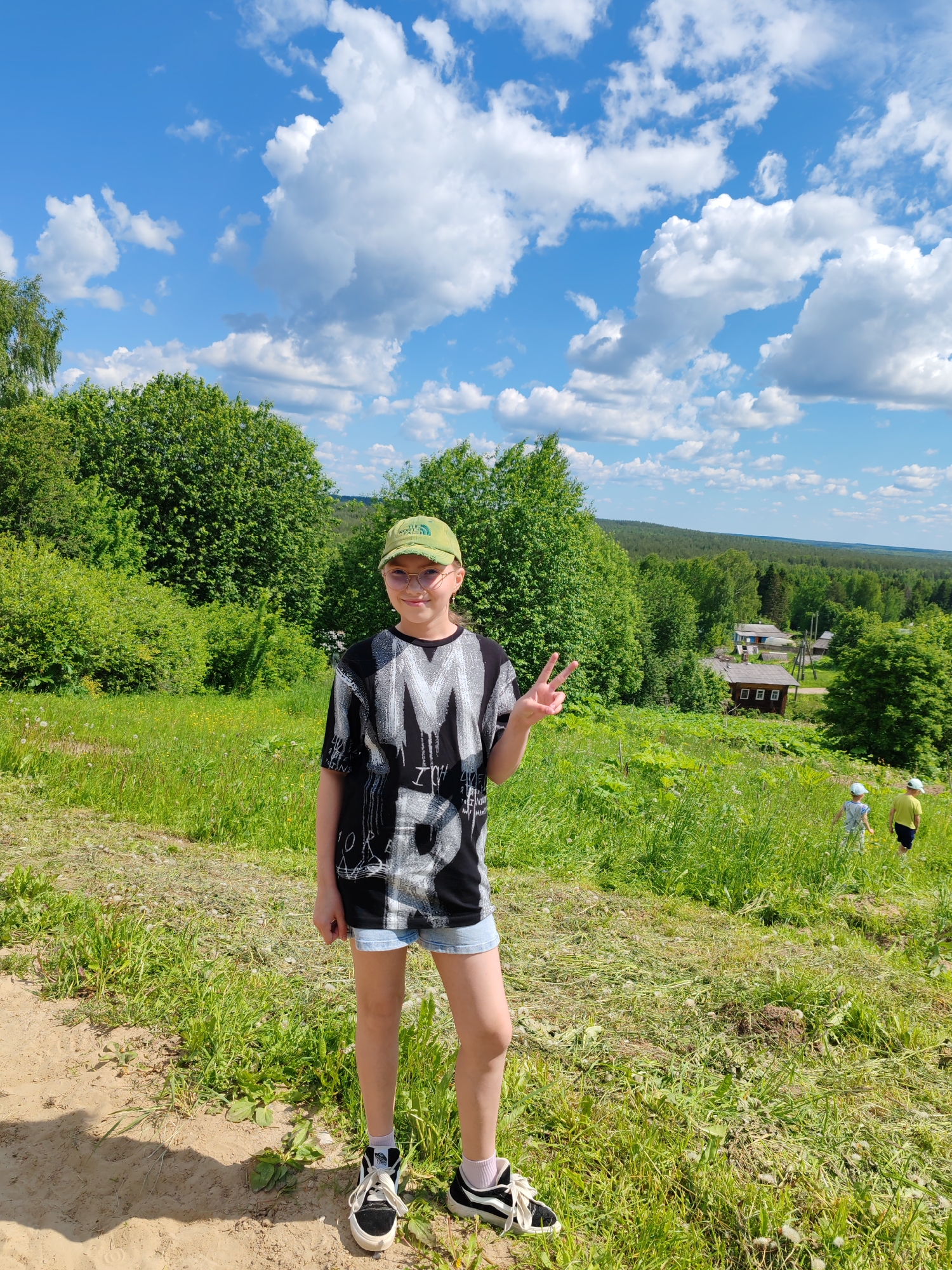 The image displays a sunny, rural landscape located in Чухлэм, Russia. A young individual, appearing to be a child, stands centrally in the foreground on an unpaved dirt path. This person wears a green baseball cap, round eyeglasses, a black and white graphic t-shirt, light blue denim shorts, and black and white high-top sneakers. Their right hand is raised, forming a peace sign. Behind the subject, the terrain slopes downwards, covered in vibrant green grass and dense deciduous trees and bushes. In the middle ground to the right, a small village cluster is visible, featuring several rustic houses nestled among the trees, one of which exhibits a blue roof element. Further down the grassy slope on the right, two additional smaller individuals, likely children, wearing hats and light-colored clothing, are observed walking away from the foreground. The distant background reveals rolling hills densely covered with forest. The sky is clear blue with numerous fluffy white cumulus clouds.