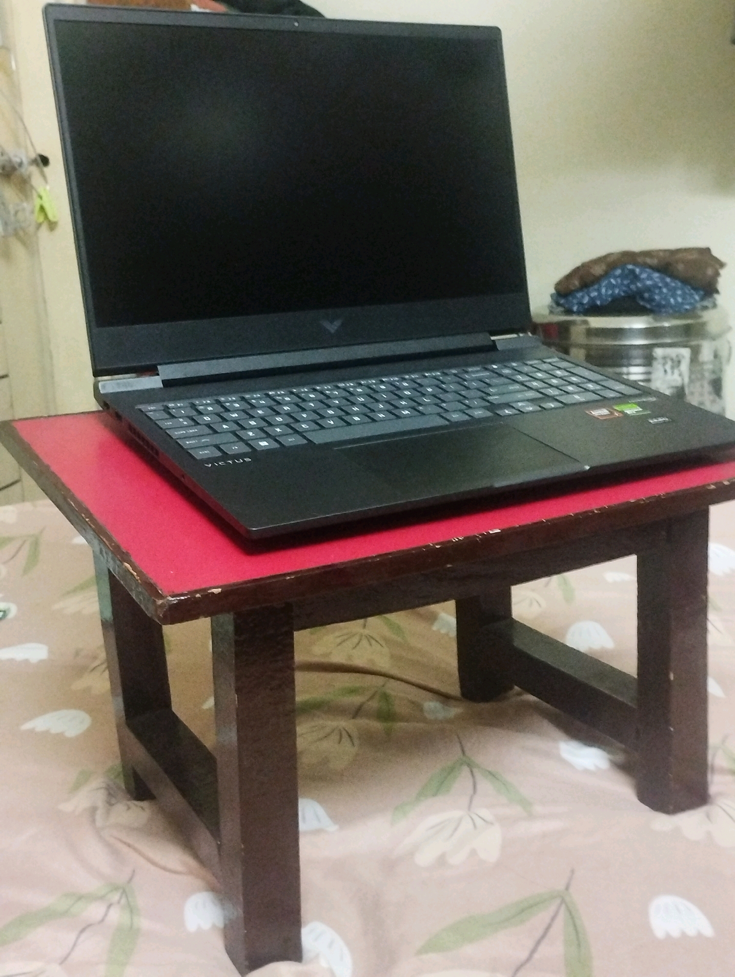 A black HP Victus laptop is positioned on a small, red-topped wooden table. The laptop is open, with its screen displaying a black image. The table is resting on a bedspread with a floral pattern featuring white flowers and green leaves. In the background, there is a metallic container and a pile of folded fabrics. The scene appears to be indoors, possibly in a residential setting in Gana, India.