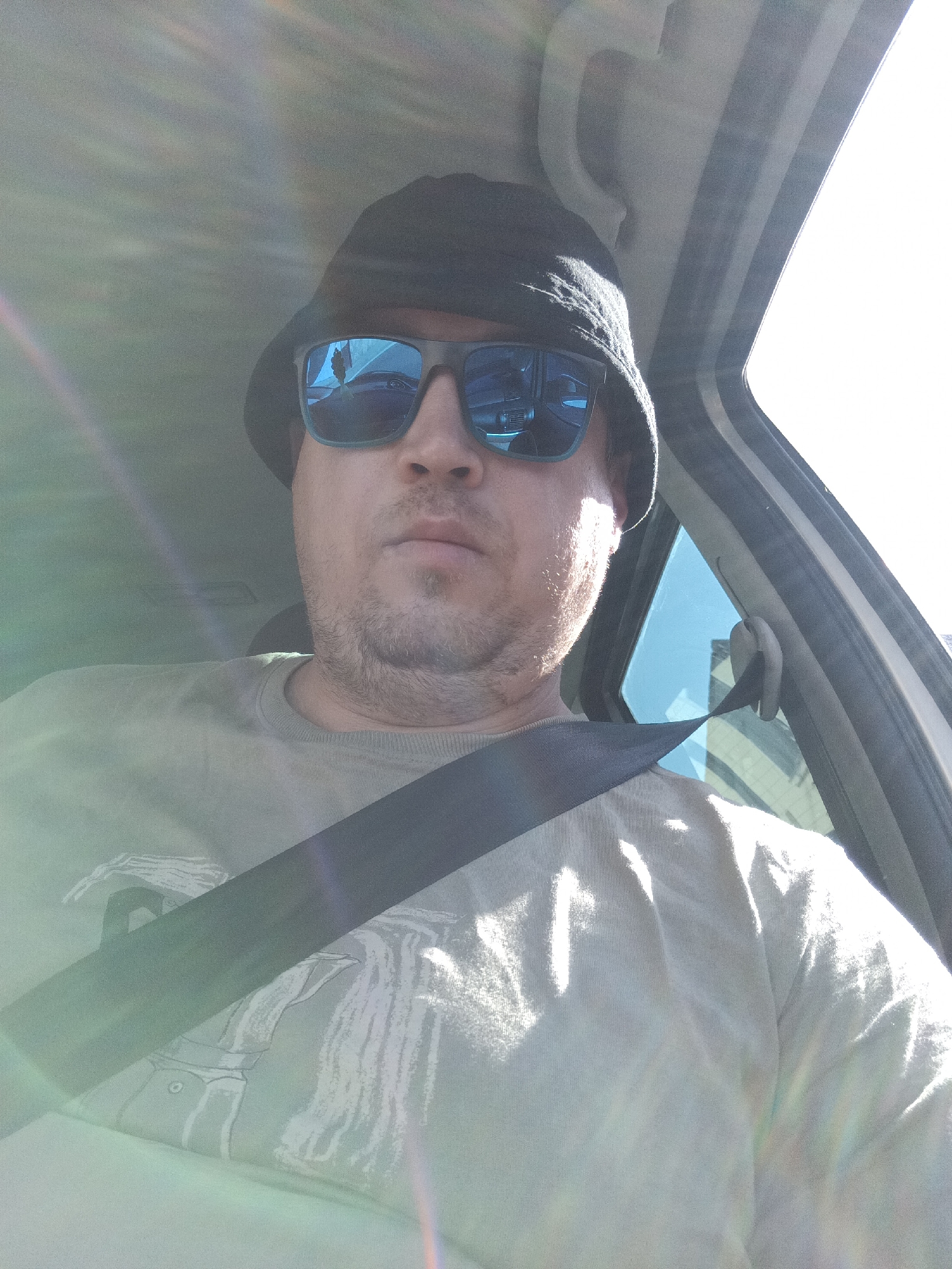 A man wearing a black bucket hat and blue mirrored sunglasses is in the driver's seat of a car. He is wearing a light-colored t-shirt with a graphic print and a black seatbelt is across his chest. The sun is shining brightly, casting lens flares on the image. The background shows a car interior with a window on the right side, revealing a glimpse of buildings and blue sky. The image appears to be a selfie taken inside the vehicle.