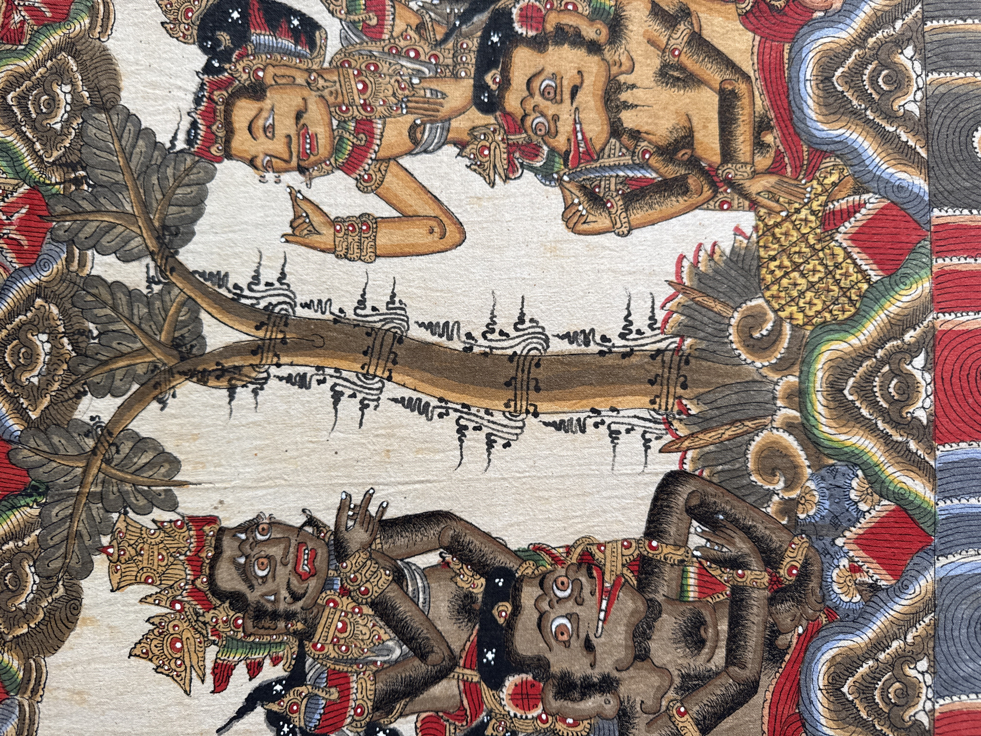 The image is a close-up of a traditional Balinese painting on fabric, depicting four figures with exaggerated features, possibly from a Wayang Kulit (shadow puppet) performance or a similar folk art tradition.
On the left, a dark-skinned figure with a grimace and elaborate golden headdress is positioned behind a lighter-skinned figure with an open mouth and wide eyes. Both wear ornate, multi-colored garments adorned with gold patterns and red, green, and black accents.
To the right, a lighter-skinned figure with a stern expression and a golden headdress is gesturing with one hand. Below and slightly to the right of this figure is another dark-skinned figure with a wide, toothy grin and bulging eyes, also adorned in similar colorful attire. A stylized tree trunk with dark, spiky branches is centrally located in the composition, separating the figures. The background is a neutral off-white, suggesting the fabric itself. The lower portion of the painting shows decorative patterns in red, blue, gray, green, and gold, forming abstract shapes.
The style of painting, with its bold outlines, vibrant colors, and stylized figures, is characteristic of Balinese art. While Kuta Selatan, Indonesia, is known for its tourism and beaches, this artwork represents a different facet of Indonesian culture, focusing on traditional storytelling and performance art. The scene itself does not provide specific environmental cues that can be definitively linked to Kuta Selatan, but rather a general representation of a cultural motif.