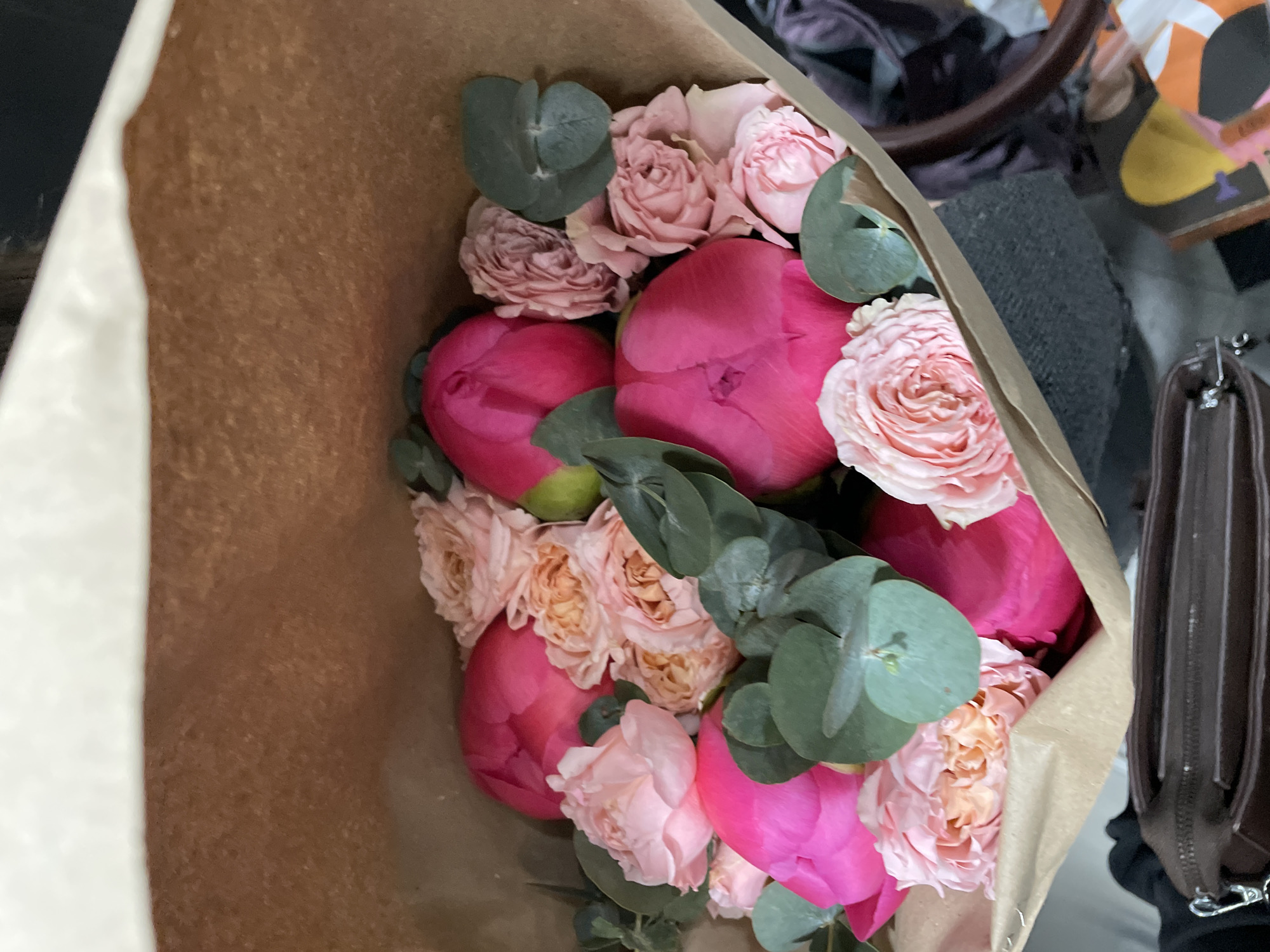 The image shows a bouquet of pink and peach-colored flowers, including peonies and roses, wrapped in brown paper. The bouquet appears to be indoors, possibly on a surface. Other objects visible include a dark bag with a brown handle, and some indeterminate items in the background. The image does not provide sufficient information to determine the location in Krasnoyarsk, Russia.