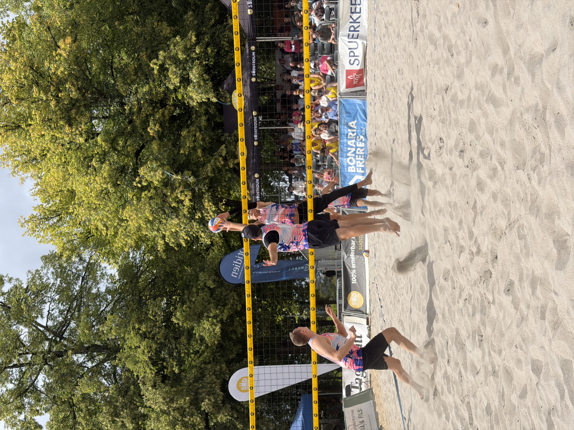 This is a photograph taken at the beach volleyball tournament in Esch-sur-Alzette, Luxembourg. The image captures a dynamic moment during a beach volleyball match. In the foreground, two male athletes are mid-air, engaged in a play over the net. One athlete on the left, wearing black shorts and a pink and white patterned tank top, is positioned lower, seemingly preparing to receive or block. The other two athletes are closer to the net and the ball. One athlete, wearing a black cap, black shorts, and a similar pink and white patterned tank top, is jumping high and reaching to hit the ball, which is visible above him. Another athlete, partially obscured, is also in the air near him, likely participating in the same play. Sand is being kicked up around the athletes' feet, indicating their vigorous movement.
In the background, a volleyball net with yellow accents divides the court. Behind the net, spectators are seated in bleachers, watching the game. A large crowd of people is visible, with some standing and others seated. The court is made of sand, and various banners and flags are displayed around the perimeter. These include banners with logos such as "SUDSTROUM," "BONARIA FRERES s.a.r.l.," and "SPUERKEESS," suggesting these are sponsors of the event. Trees with green foliage form a backdrop to the scene, indicating an outdoor setting. The sky is partly cloudy. The overall impression is one of an active sporting event taking place on a sunny day.