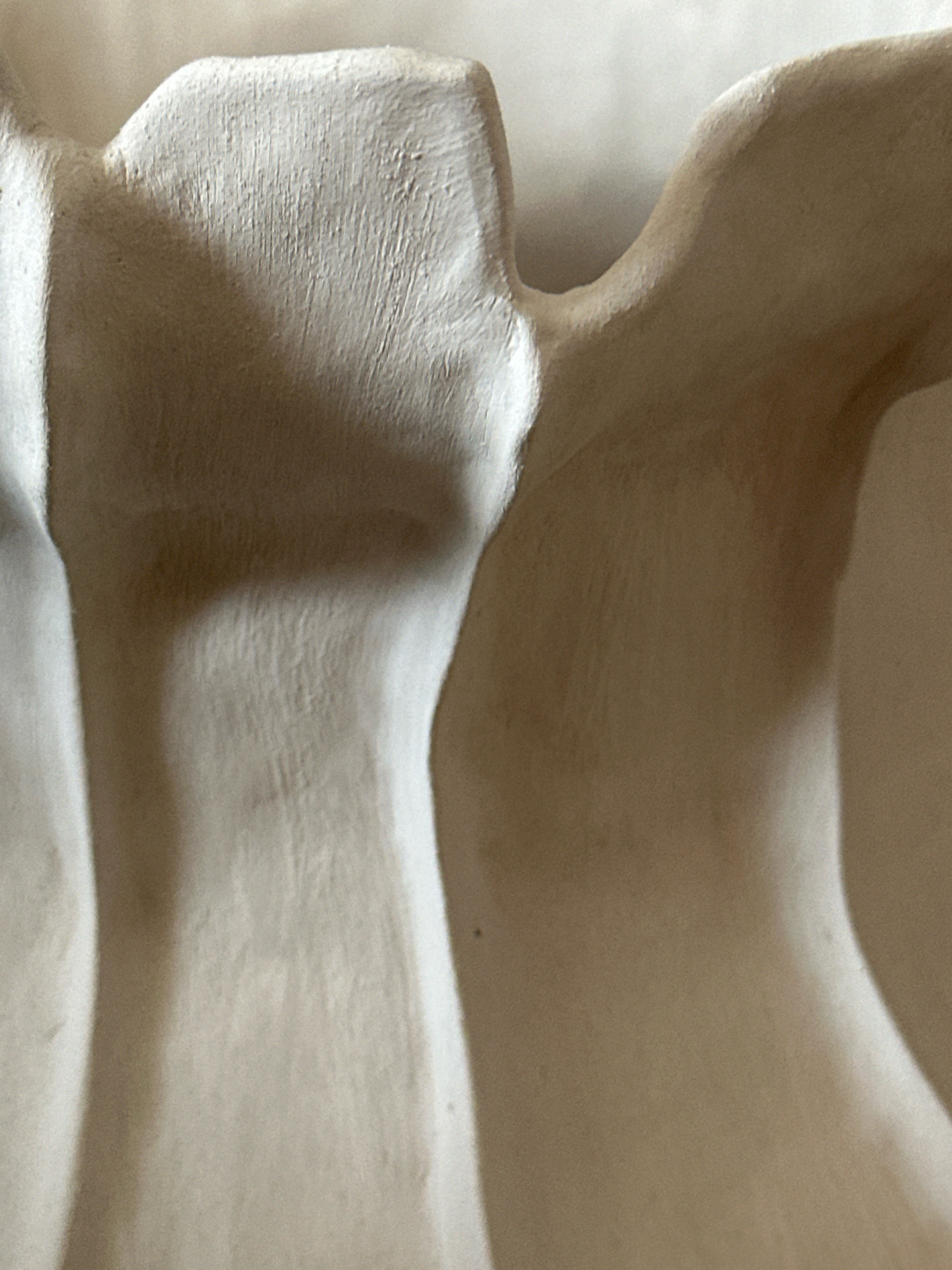 The image is a close-up, abstract shot of a textured, light-colored object, possibly ceramic or plaster. The lighting creates deep shadows and highlights, emphasizing the curves and ridges of the material. There are visible vertical striations and subtle imperfections on the surface, suggesting it might be handmade. The overall impression is one of organic form and tactile quality. This object is not identifiable as being located in Nevinnomyssk, Russia, or any specific urban environment.