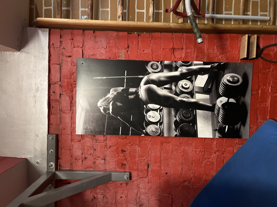 A workout space featuring a red brick wall. A black and white poster of a woman lifting weights hangs on the wall above a blue exercise mat. A gray pull-up bar is mounted to the left side of the wall. A wooden rod and metal equipment are partially visible at the top of the frame.