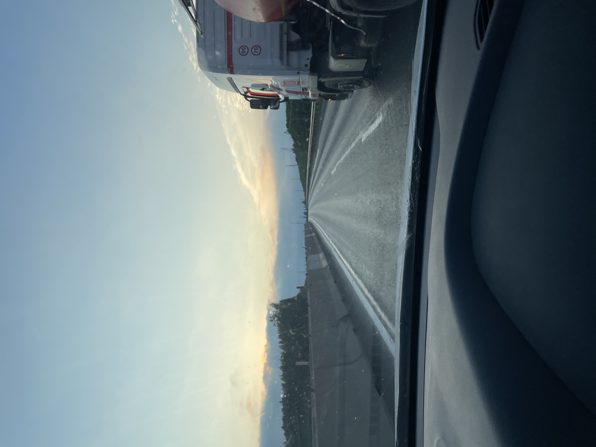 The image is taken from inside a vehicle, looking out at a highway at dusk. The sky is a gradient of light blue and yellow, with wispy clouds. The road ahead is wet, reflecting the light. To the right of the road is a white semi-truck with red trim. On the side of the truck, there are circular signs indicating speed limits of 90 and 70. The truck is partially obscured by the edge of the frame. On the left side of the road, there is a concrete barrier, and beyond that, a line of trees and hills. The interior of the vehicle, specifically the dashboard and windshield, are visible in the foreground, indicating the perspective is from the driver's seat. The location is identified as Novozhilovo, Russia.