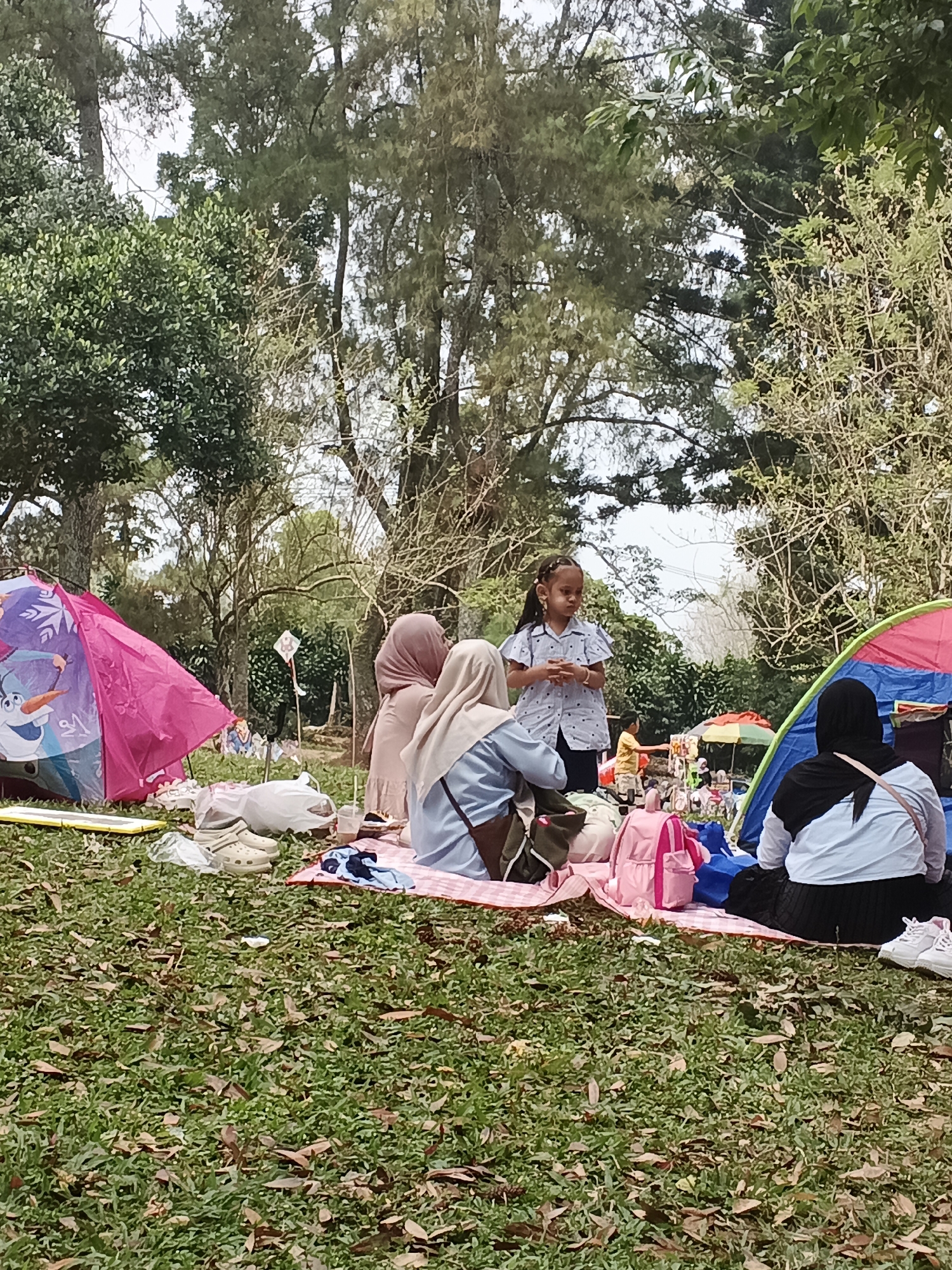 In the city of Perbawati, Indonesia, a group of people are enjoying a picnic in a grassy, wooded area. A pink tent with a cartoon character is set up on the left, and a colorful tent is on the right. Several individuals, including women wearing hijabs and a young girl, are gathered around picnic blankets spread on the ground. Various items, such as bags, a drink, and shoes, are scattered around the blankets. In the background, trees and foliage create a natural, outdoor setting.