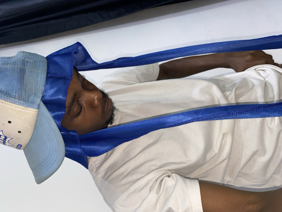 A man is seen sleeping. He is wearing a light blue hat and a blue satin wrap. He is lying on his back, wearing a white t-shirt. His eyes are closed. His hand is on the right side of his body. The background is white and black, with a shadow in the top corner.