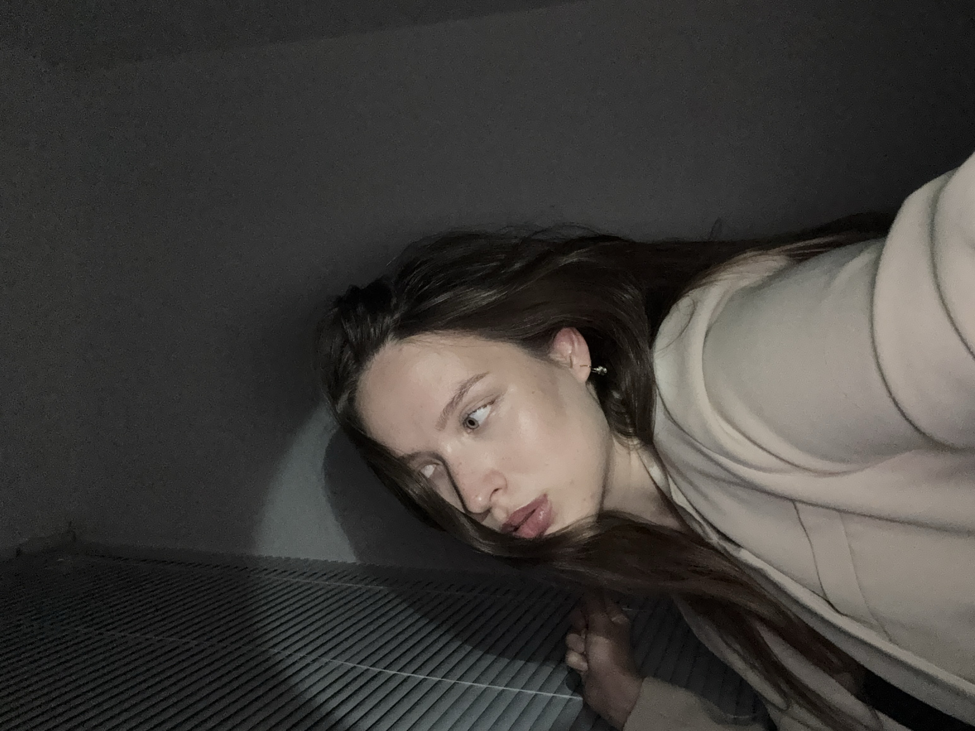 The image displays a young adult female captured in a close-up, indoor setting with subdued lighting. She has long, dark brown hair, styled with a side part, flowing over her shoulders. Her complexion is fair, and she possesses light-colored eyes, directed upwards and to the viewer's left. Her mouth is slightly agape, suggesting a pensive or observant expression. A small, silver-toned dangle earring is visible in her right earlobe. She is dressed in a light beige or cream-colored blazer or jacket, with a darker garment visible underneath. Her left hand is positioned near a window area.
To the left of the subject, a set of dark, horizontal window blinds or vertical slats covers a window. The primary light source appears to emanate from the left, illuminating her face and the blinds, while casting subtle shadows on the right side of her face and the adjacent wall. The background on the right side of the image consists of a plain, dark grey wall, and the ceiling above is also dark.
The image contains no identifiable visual markers, such as specific landmarks, architecture, or geographic features, that would permit a definitive identification of the location as the city of Chaikovsky, Russia.
