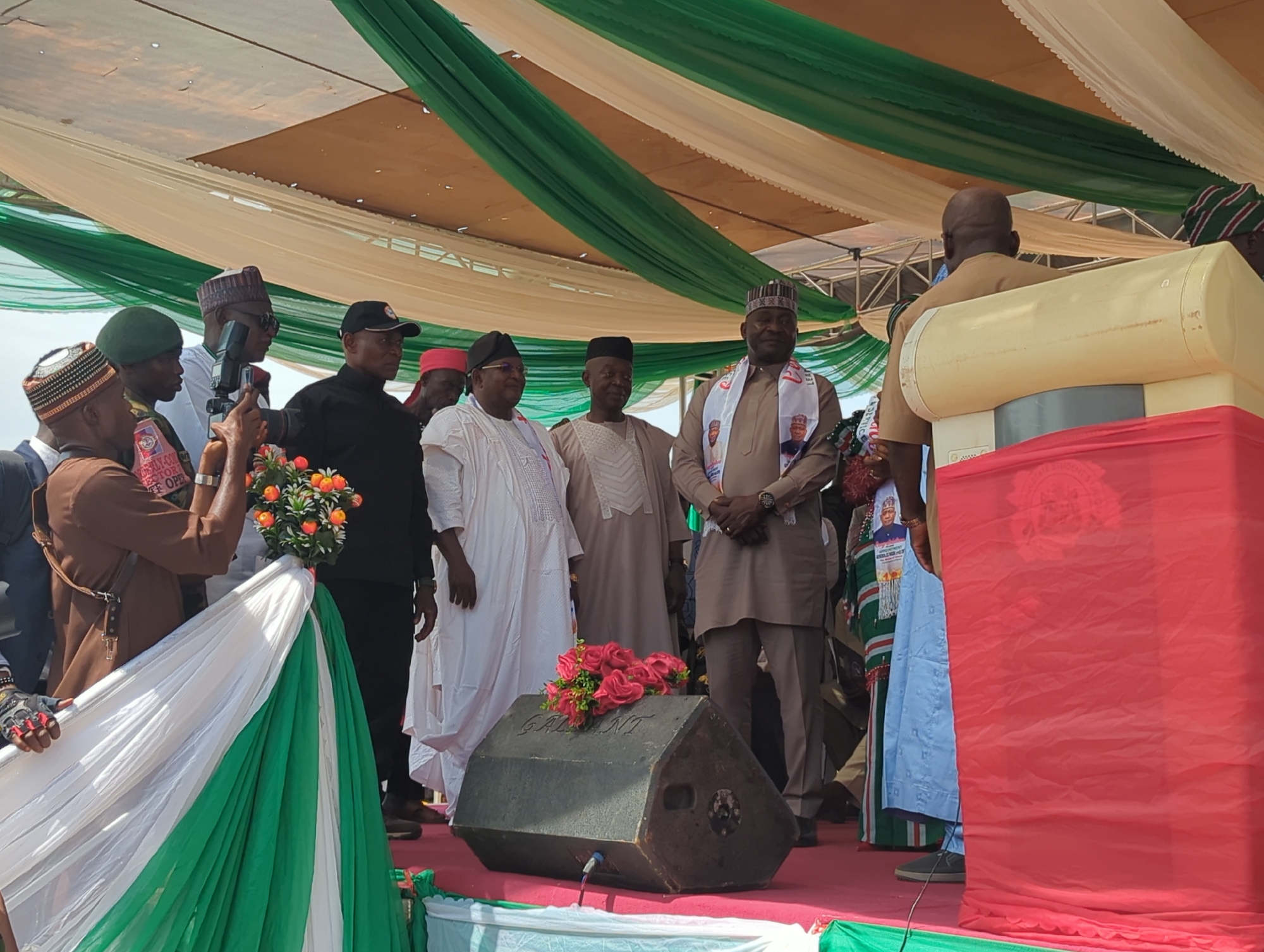 A group of men are on an outdoor stage in Takau, Nigeria. The stage is decorated with the colors of the Nigerian flag (green and white) and has beige and green drapes hanging overhead. Five men are standing together, facing the camera. To the left, a man is taking a picture of them with a camera. In front of the men is a loudspeaker decorated with roses, and a podium covered in red cloth. The man at the podium is giving a speech. The overall atmosphere appears to be formal and celebratory.