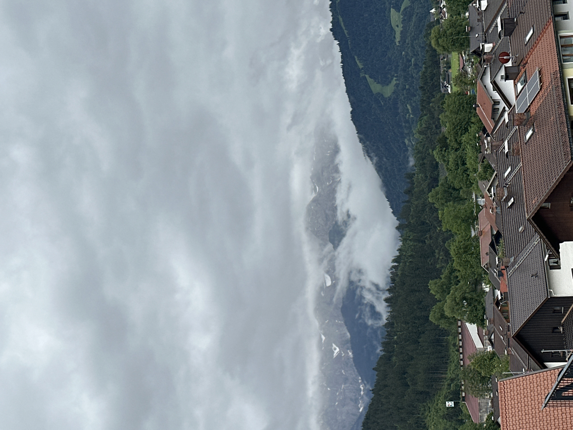 The image shows a cloudy, overcast sky over a mountainous landscape with a village in the foreground. The mountains are covered with dark green coniferous trees, and some higher peaks are visible, with patches of snow or rock. Clouds are obscuring the upper portions of the mountains, creating a hazy effect. A ski slope is visible on one of the mountain faces.
In the middle ground, a dense forest of trees lines the base of the mountains. Below the forest, a cluster of buildings forming a village is visible. These buildings have pitched roofs covered with tiles in various shades of red and brown. Some buildings have visible chimneys, solar panels, and skylights. There are no people visible in the image.
The foreground shows the tops of more buildings with tiled roofs. A dark metal railing is partially visible in the lower left corner. The overall scene depicts a typical alpine village in Germany, likely in a mountainous region such as Garmisch-Partenkirchen, given the presence of ski slopes and the architectural style of the buildings.