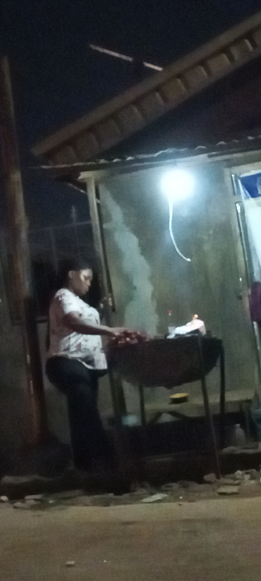 At Sabon Tasha, Nigeria at night, a woman wearing a light-colored patterned shirt and dark pants stands beside a charcoal grill. The grill contains food being cooked and is producing smoke. The woman appears to be tending to the grill. Behind her is a building with an overhanging roof supported by wooden posts. An electric light illuminates the immediate area. The ground appears to be paved or covered with dirt.