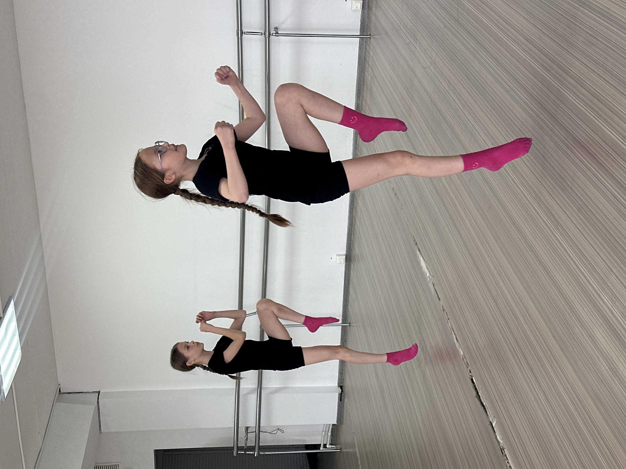 The image depicts a young female in a dance studio. The location, Votkinsk, Russia, is unknown, but the interior setting suggests a ballet or dance school. She is wearing a black leotard and bright pink socks with a smiley face on the side. Her hair is long and braided. She is reflected in a mirror. In front of the mirror is a horizontal metal bar which likely serves as a ballet barre. She appears to be in a preparatory pose or exercise, with one knee up and hands in fists. The room is well-lit, with artificial lighting coming from above. The floor is a light-colored wood material. There is a radiator on the wall.