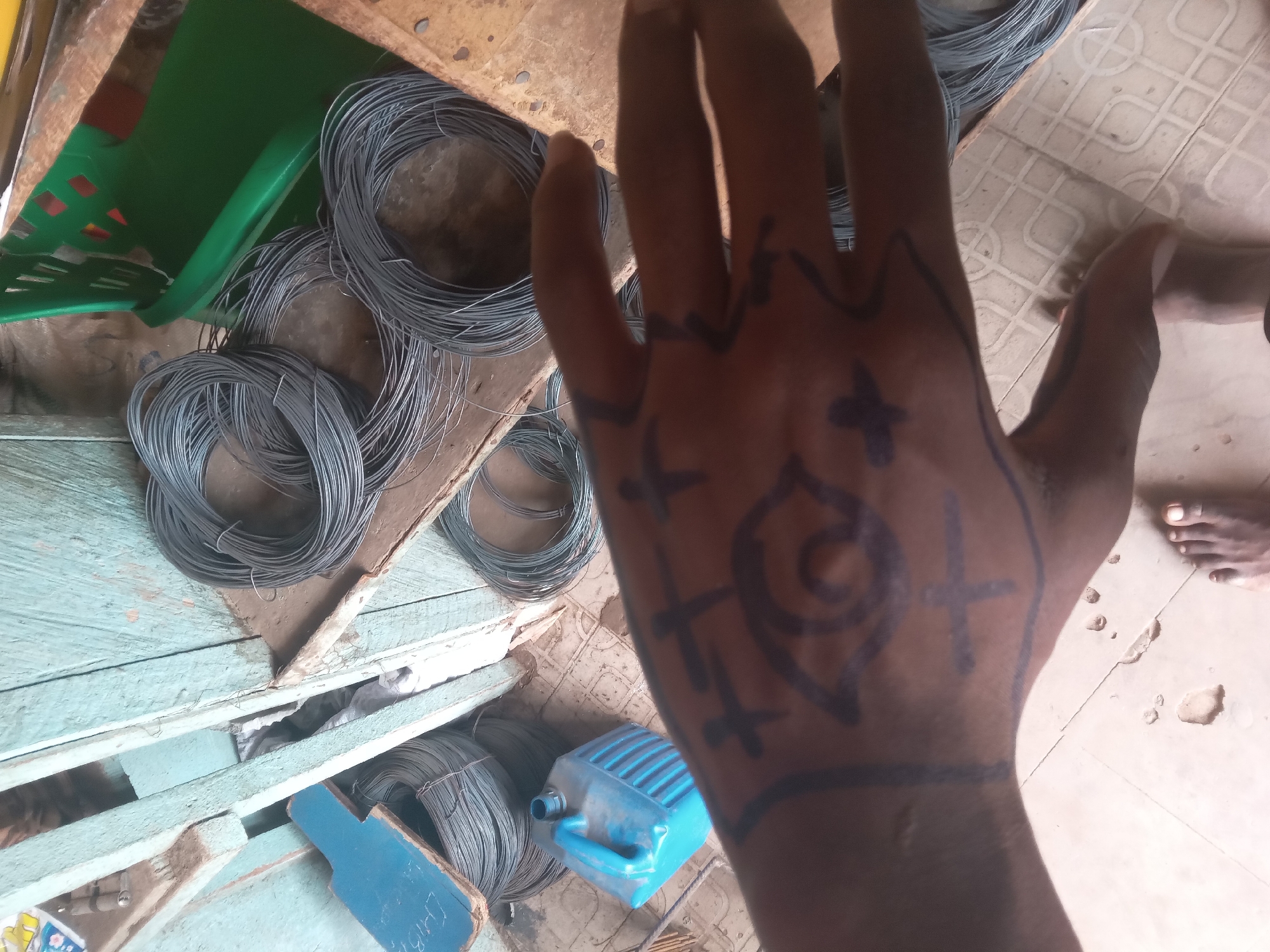 A close-up, high-angle shot depicts a hand with drawings in black ink against a background of wire coils, plastic containers, and wooden surfaces. The hand, with brown skin, displays various symbols, including crosses and a spiral-like design. The background reveals multiple coils of thin, grey wire, suggesting a construction or metalworking context. There is a green plastic container and a blue plastic container present. The wooden surfaces appear aged and possibly painted with blue paint. Parts of a foot is visible in the upper right. The scene might be located in Onitsha, Nigeria, given the objects and colors.