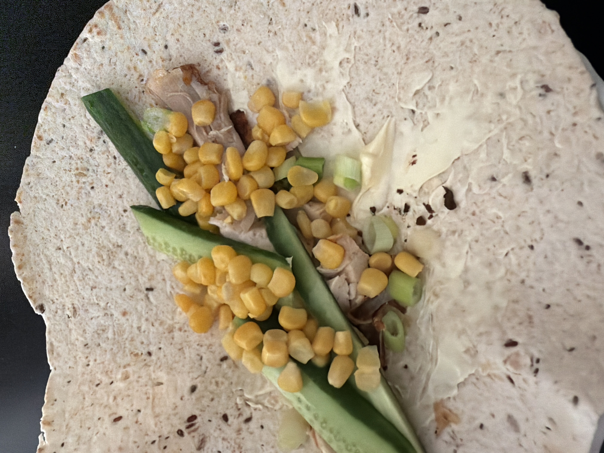 A close-up, eye-level shot shows a partially assembled wrap on a speckled flatbread. The wrap filling consists of shredded cooked chicken, bright yellow corn kernels, thin slices of cucumber, and chopped green onions. A creamy white sauce, likely mayonnaise, is spread on the flatbread underneath the fillings. The background is dark and out of focus, suggesting the wrap is on a dark surface. The scene is indoors, with no indication of the specific location within Tersefanou, Cyprus.
