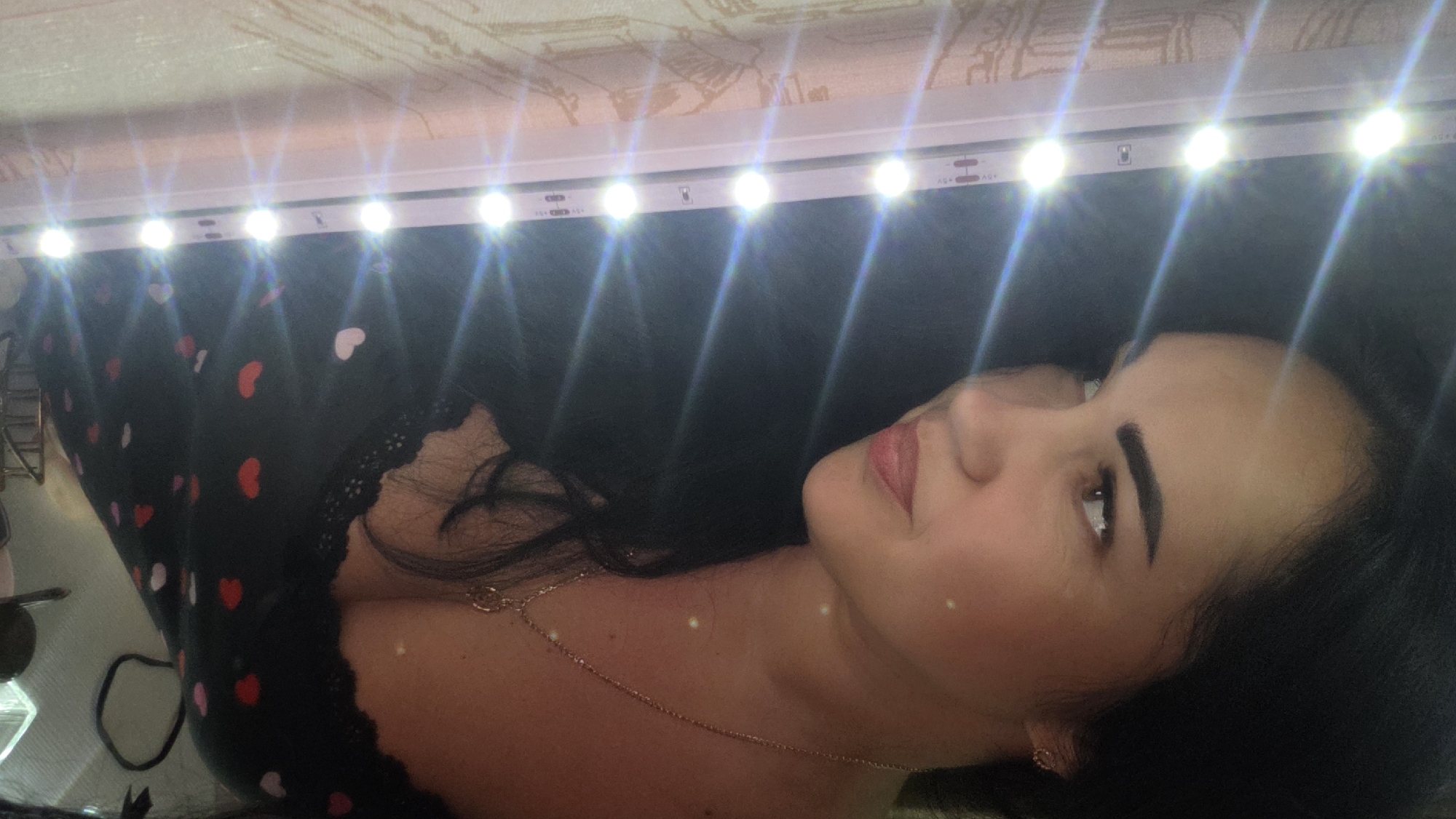 A medium shot captures a woman with dark hair, looking to her left with a slight smile. She wears a black top with a pattern of red and pink hearts and a gold necklace. To her left is a strip of bright LED lights. The background is dimly lit and appears to be indoors.