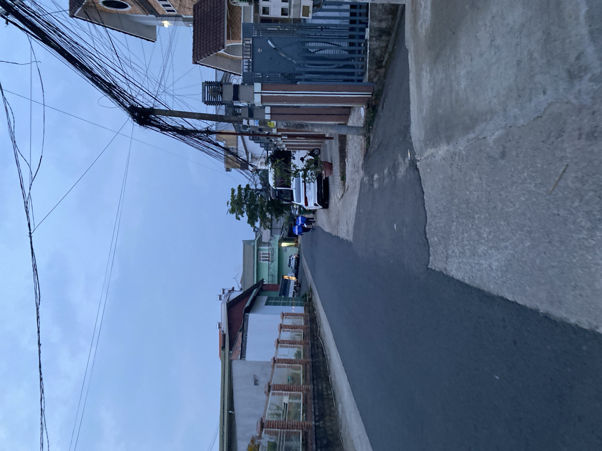 The image depicts a street in Lâm Viên - Đà Lạt, Vietnam. The street is lined with houses and power lines. The sky is overcast. A white van is parked along the road. There are some people walking in the distance.