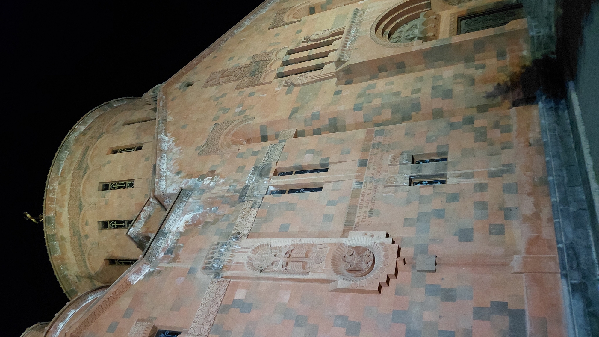 The image captures a nighttime view of a large, intricately decorated stone building, likely a church or cathedral, in Abovyan, Armenia. The structure is primarily constructed of reddish-orange and gray square stones, giving it a textured and historical appearance.
The facade features numerous decorative elements carved into the stone. These include ornate cross-like designs, circular motifs, and bands of what appear to be Armenian inscriptions running vertically and horizontally. Several small, arched windows with decorative grilles are visible, some appearing to be illuminated from within. A prominent curved section, likely part of a dome or apse, is visible on the left side of the image, also adorned with carved patterns and a series of windows. The architecture suggests a strong Armenian cultural influence.
The lighting is artificial, casting shadows and highlighting the relief carvings. The dark sky indicates it is nighttime. There are no people visible in the frame. The overall impression is one of grandeur, historical significance, and solemnity, fitting for a religious or important civic building. The weathered appearance of the stone suggests the structure is old and has stood for a considerable time.